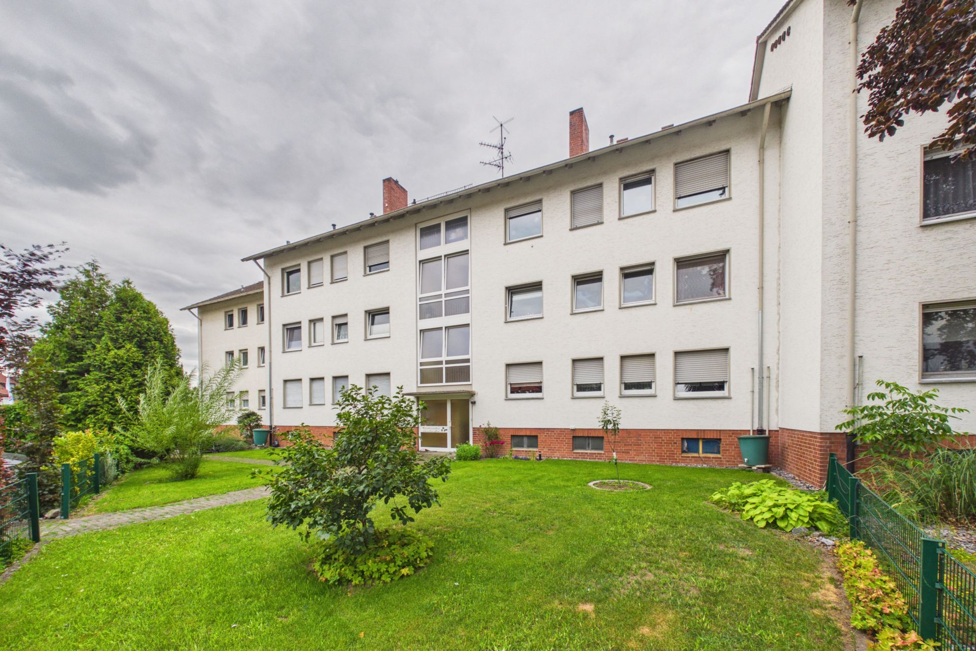 Dreistöckiges Wohnhaus mit weißer Fassade und grünem Vorgarten, Gehweg führt zum Eingang.