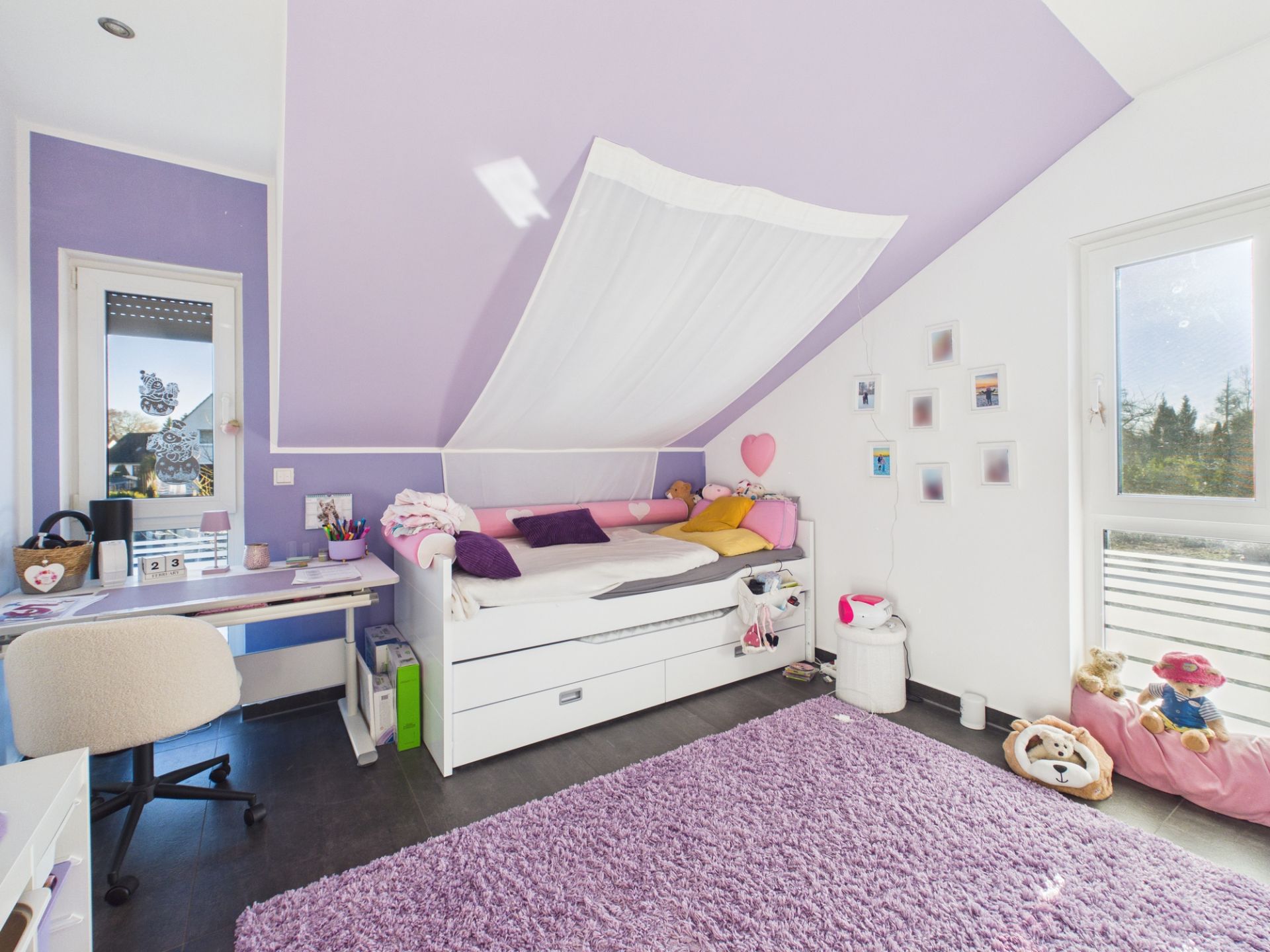 Kinderzimmer mit lila Wänden, Schreibtisch, Bett, Teppich und Fenstern.