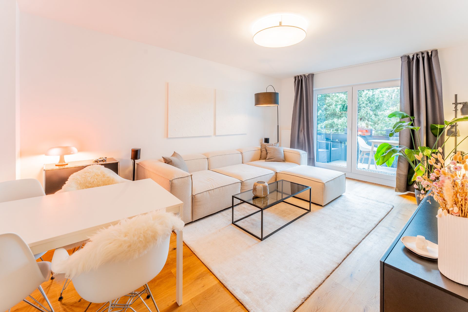 Helles Wohnzimmer mit beigem Sofa, Glastisch, Essbereich und großen Fenstern zum Balkon.
