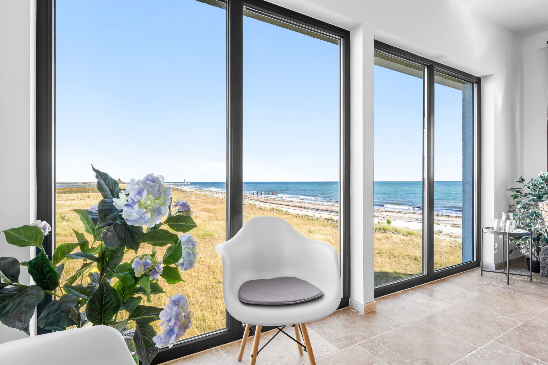 Heller Raum mit großem Fenster, Blick aufs Meer, Blumen und weißem Stuhl im Vordergrund.