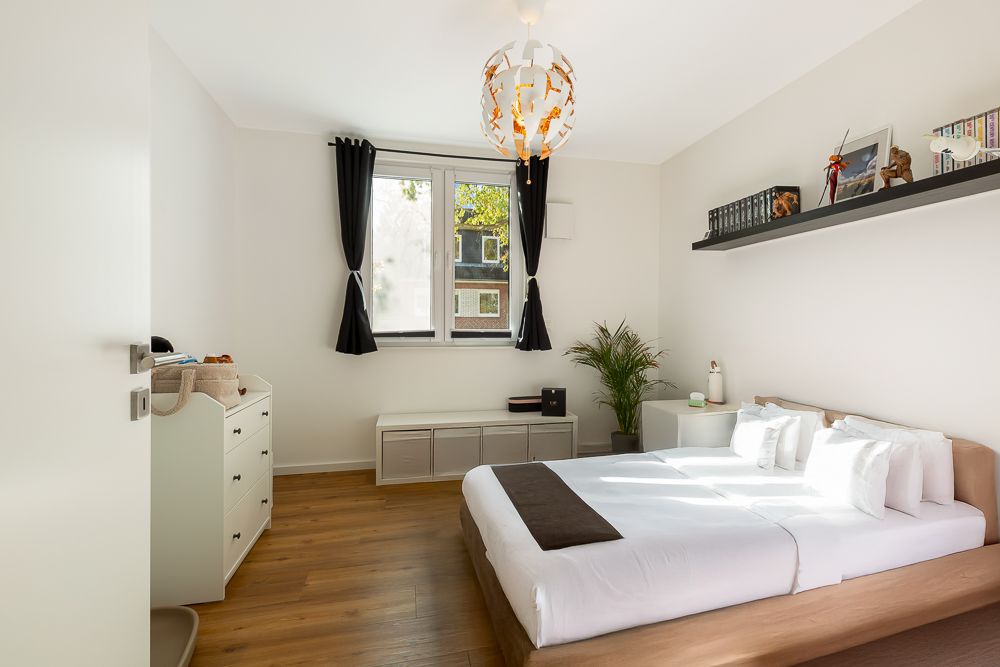 Helles Schlafzimmer mit Doppelbett, Holzboden, Fenster, Kommode und Regal über dem Bett.