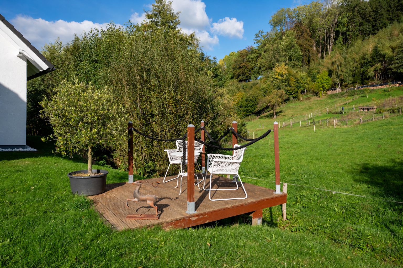 Kleine Holzterrasse mit zwei weißen Stühlen im Garten, daneben ein Baum im Topf.