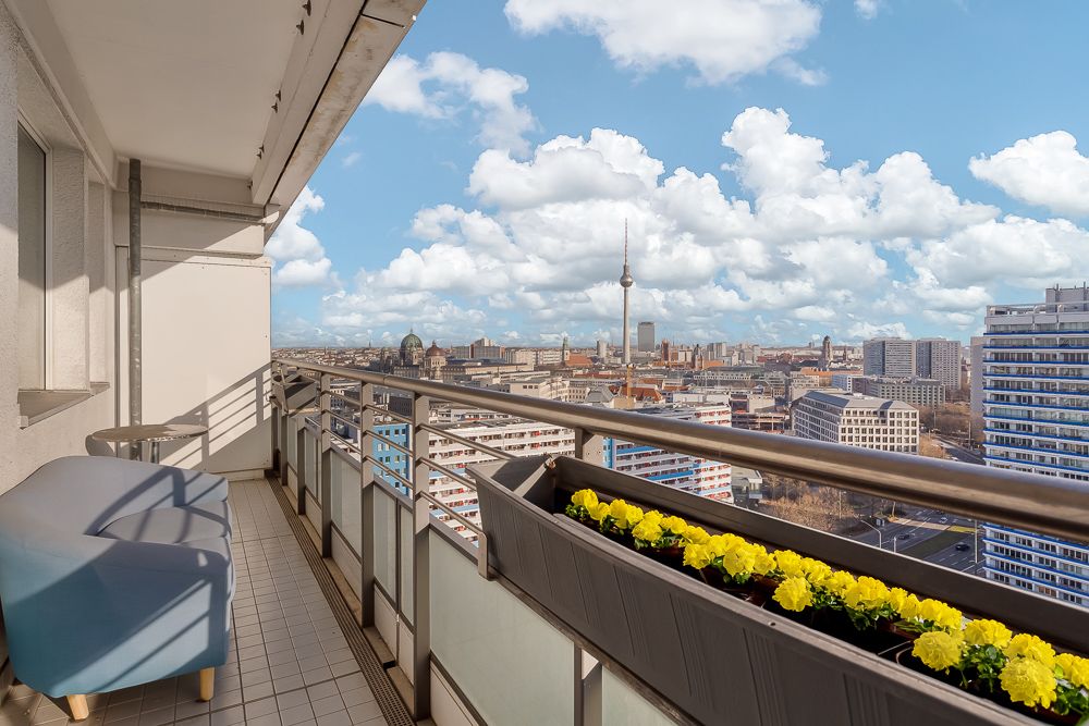 Balkon mit Blumen, Sitzbank und Blick auf eine Stadt mit Fernsehturm und Hochhäusern.
