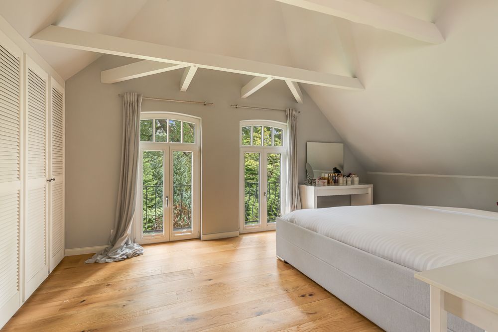Helles Schlafzimmer mit Doppelbett, Holzfußboden, zwei Fenstern und Schminktisch unter schrägem Dach.