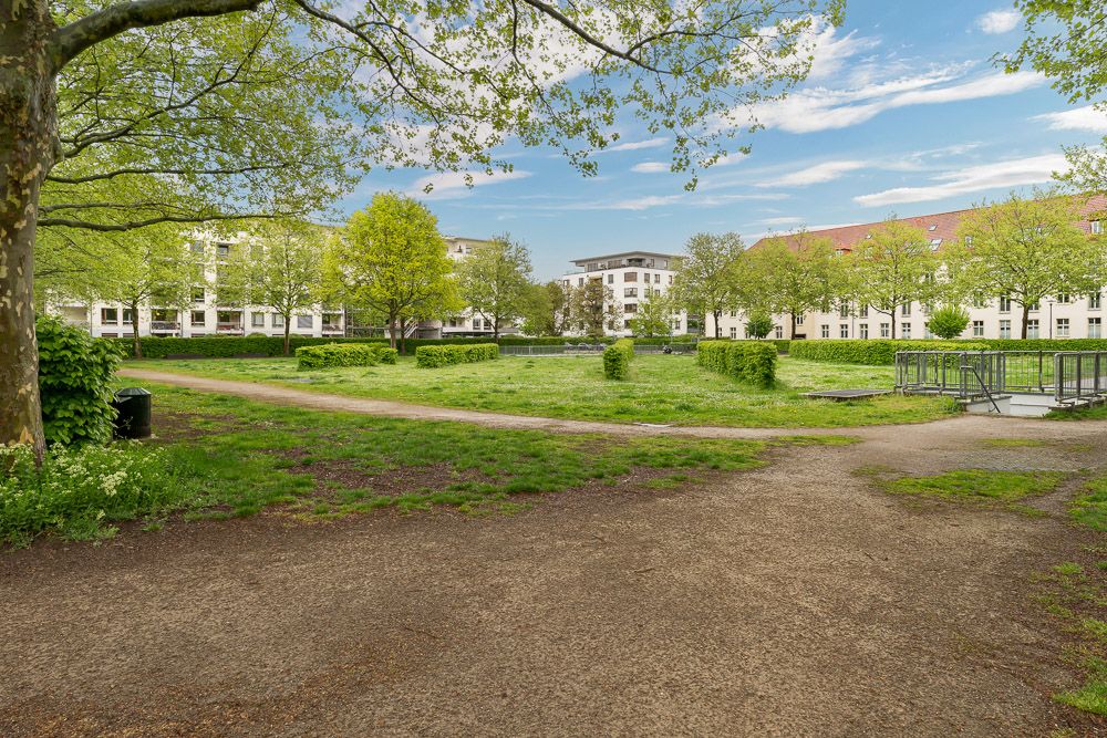 Grüner Innenhof mit Wegen, Bäumen und umgebenden Wohngebäuden bei blauem Himmel.