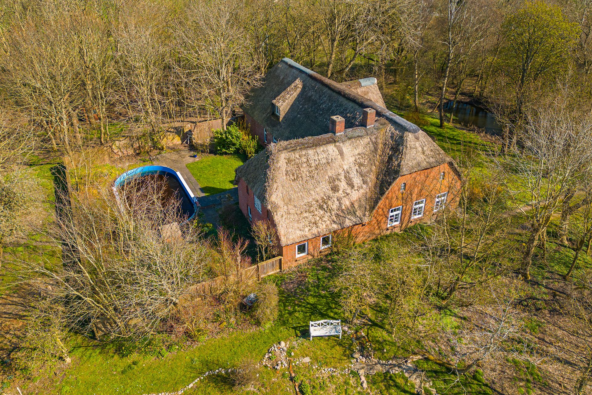 Luftaufnahme eines Backsteinhauses mit Reetdach, Garten und ovalem Pool, umgeben von Bäumen.