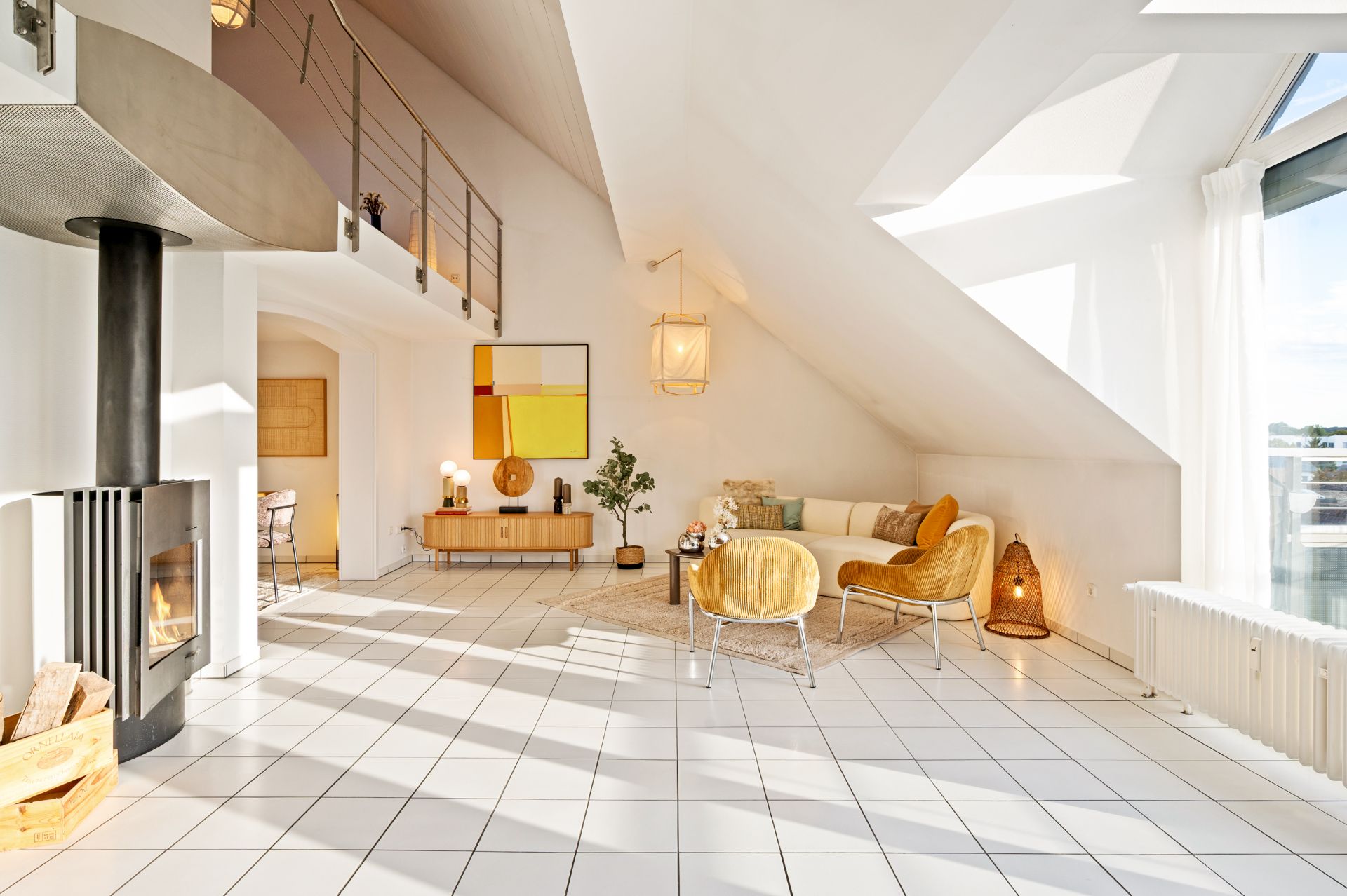 Helles Wohnzimmer mit Kamin, Sesseln, Sofa, großen Fenstern und weißem Fliesenboden.