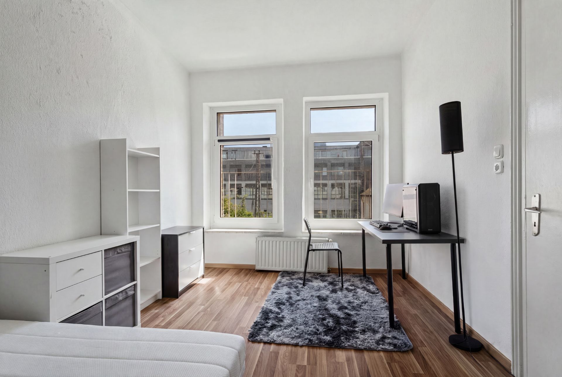 Helles Zimmer mit Schreibtisch, Regal, Stuhl, Teppich und zwei großen Fenstern.