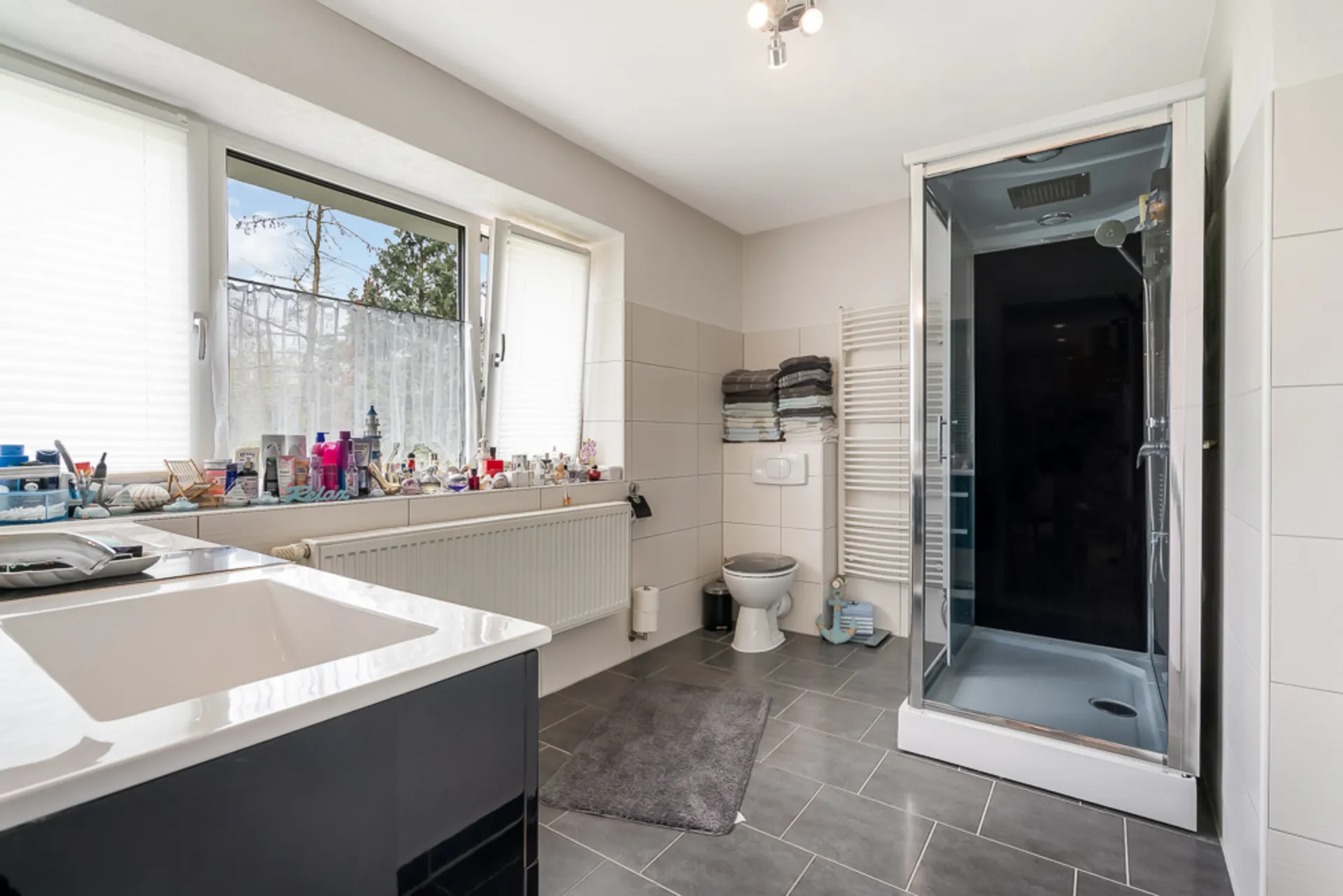 Modernes Badezimmer mit Dusche, Waschbecken, WC und großen Fenstern, viele Pflegeprodukte auf dem Fenstersims.
