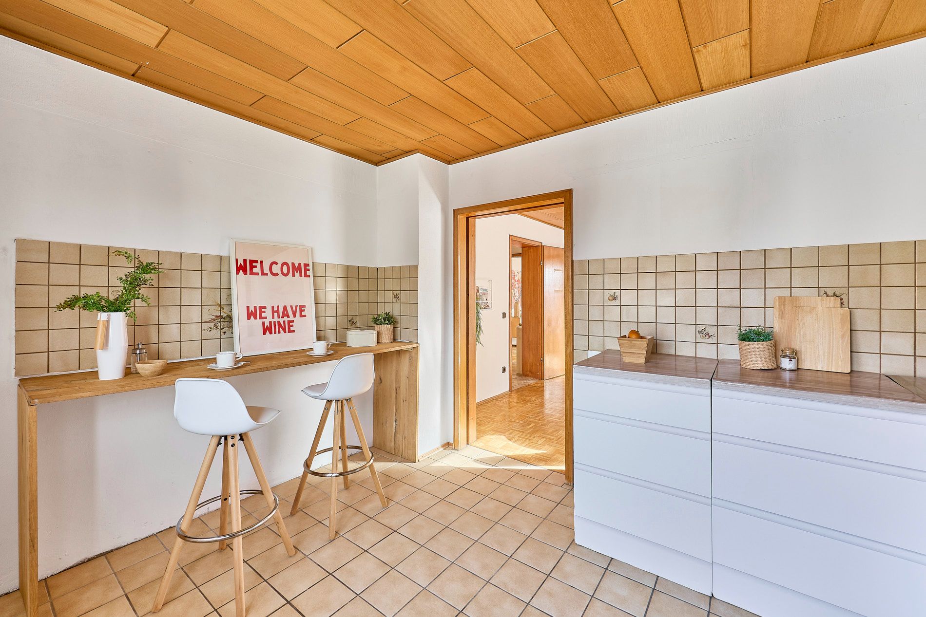Helle Küche mit Holztisch, zwei weißen Hockern und weißen Schränken, gefliestem Boden und Pflanzen.