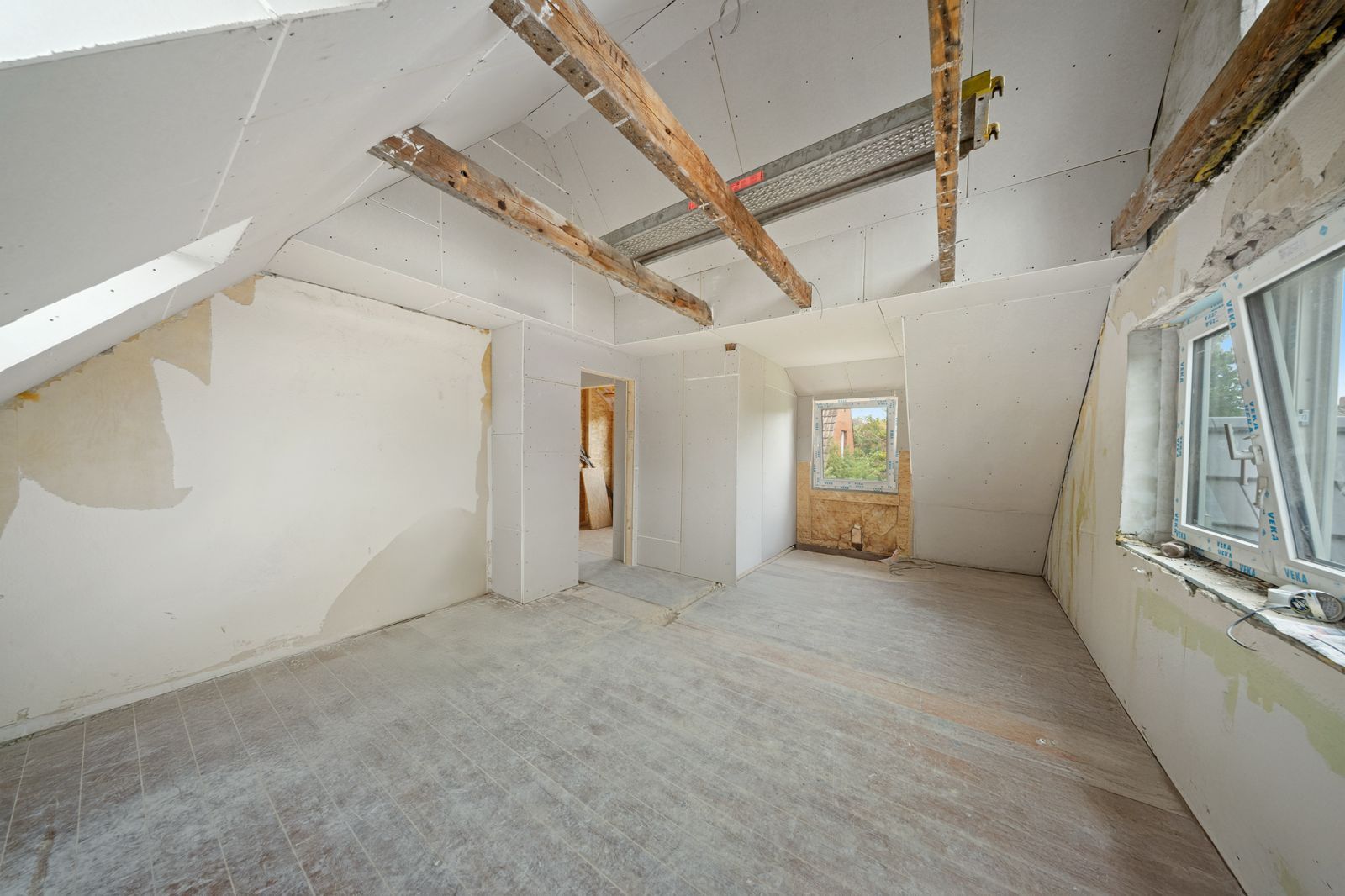 Helles Dachzimmer im Rohbau mit sichtbaren Holzbalken und zwei Fenstern.