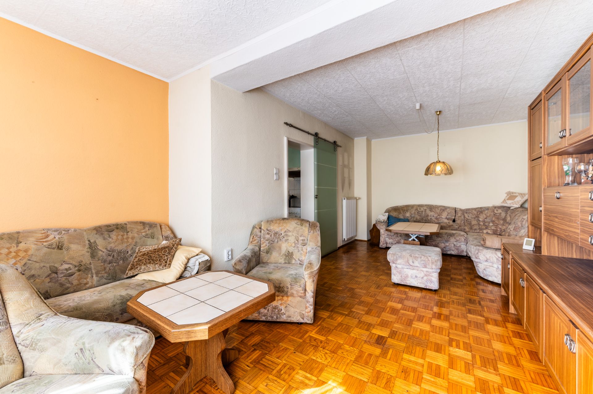 Helles Wohnzimmer mit Polstermöbeln, Holztisch, Schrankwand und Parkettboden.