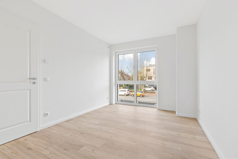 Helles, leeres Zimmer mit Holzfußboden und großem Fenster zur Straße.