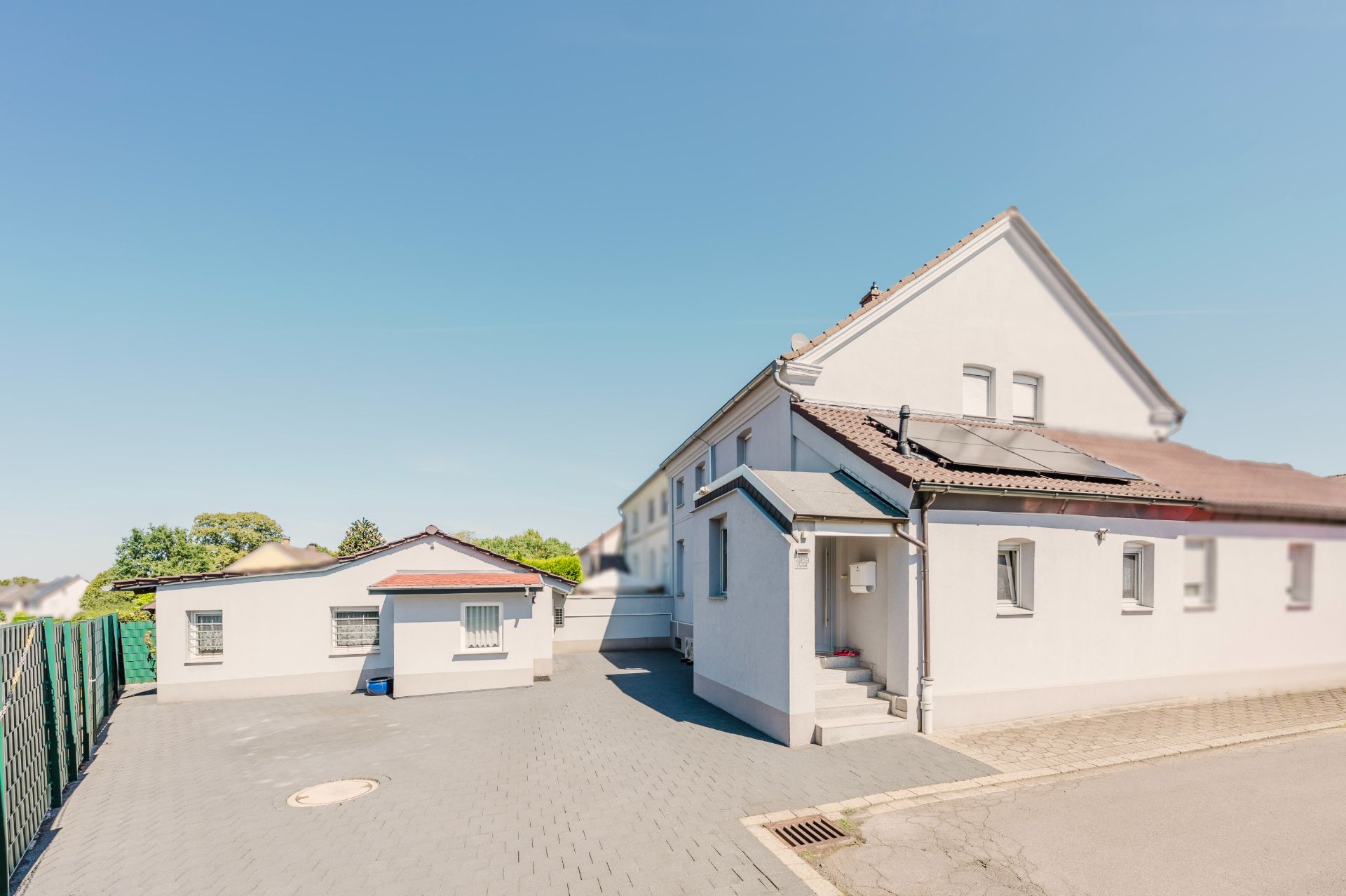 Gepflasterter Hof mit zwei weißen Wohngebäuden, einem Zaun und blauem Himmel.