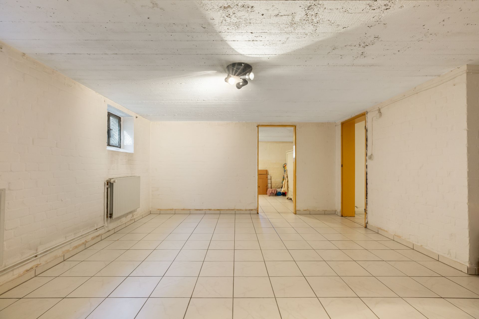 Heller, leerer Kellerraum mit Fliesenboden, weiß gestrichenen Wänden und einem kleinen Fenster.