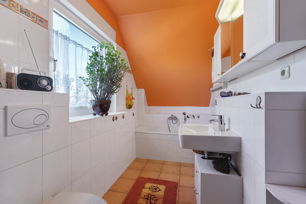 Helles Badezimmer mit oranger Wand, Fenster, Pflanze, Waschbecken, Badewanne und weißen Fliesen.