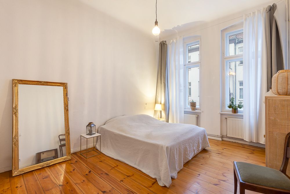 Helles Schlafzimmer mit Holzboden, Doppelbett, großem Spiegel und zwei Fenstern mit weißen Vorhängen.