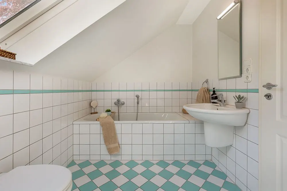 Helles Badezimmer mit Badewanne, Waschbecken, Spiegel und weißen Fliesen unter einer Dachschräge.