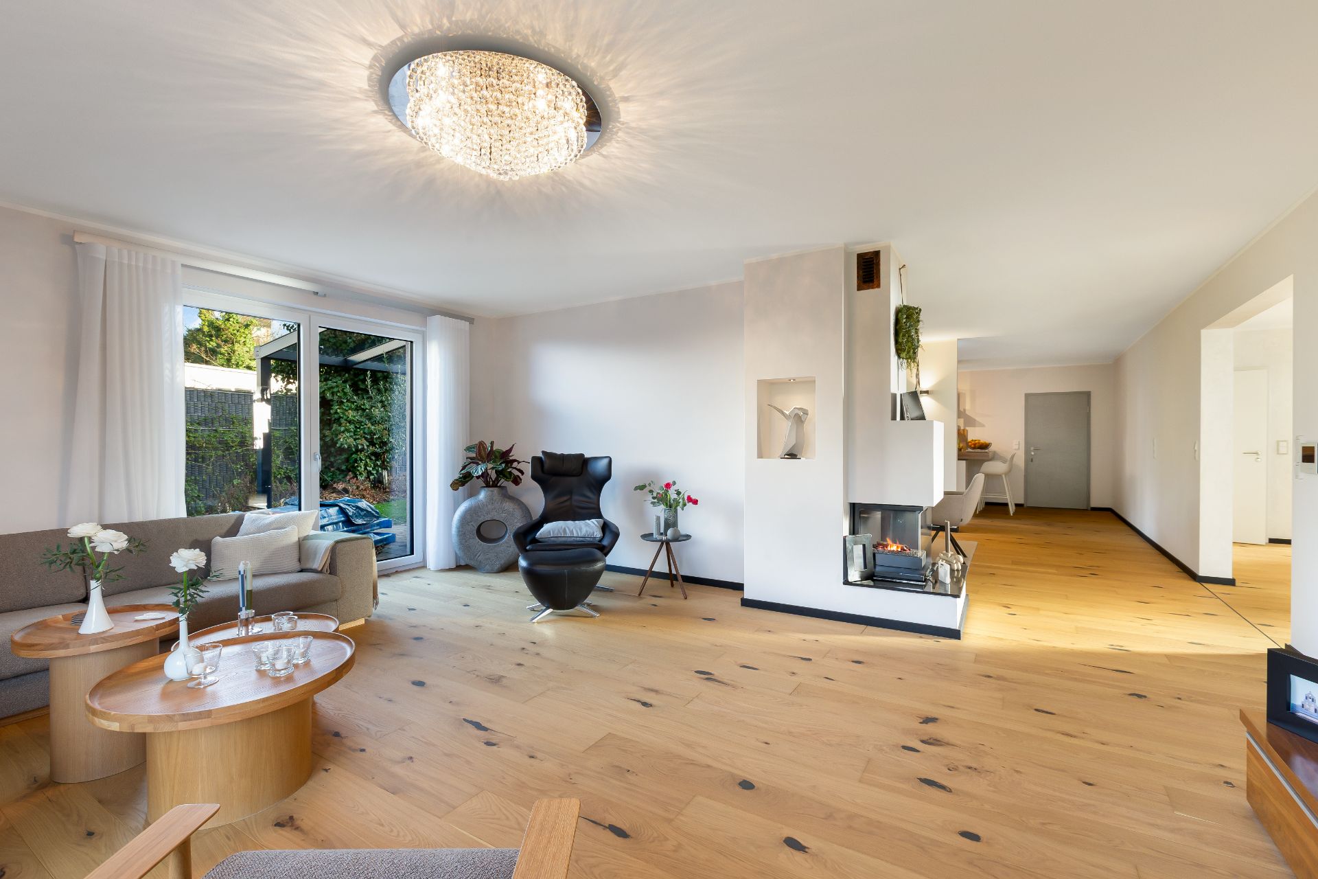 Helles Wohnzimmer mit Holzfußboden, Kamin, Sesseln, Couchtischen und großem Fenster zur Terrasse.