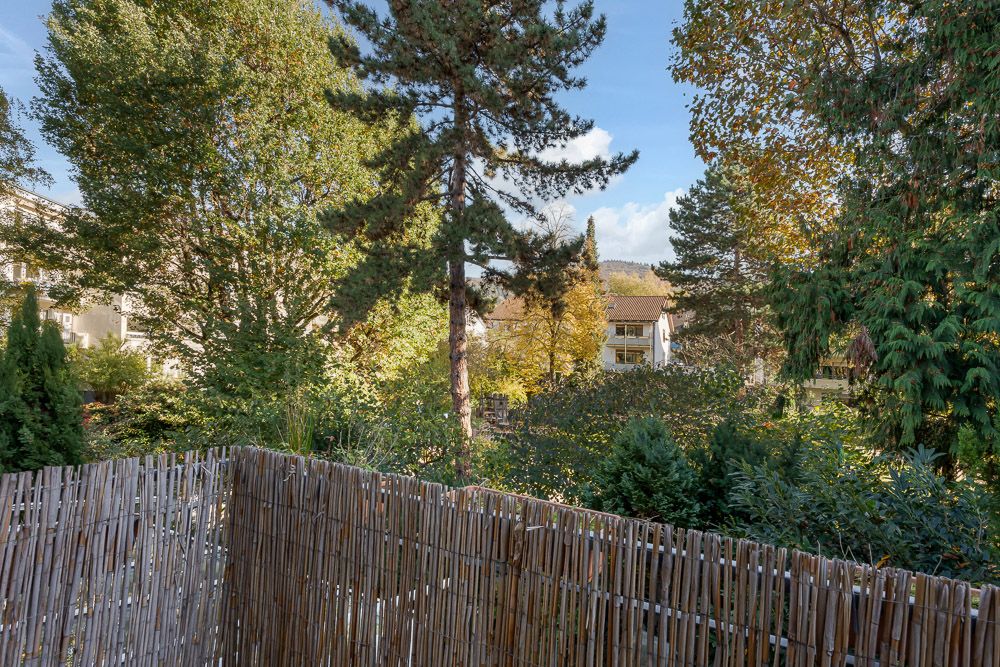 Blick von einem Balkon mit Bambuszaun auf viele Bäume und ein Wohnhaus im Hintergrund.
