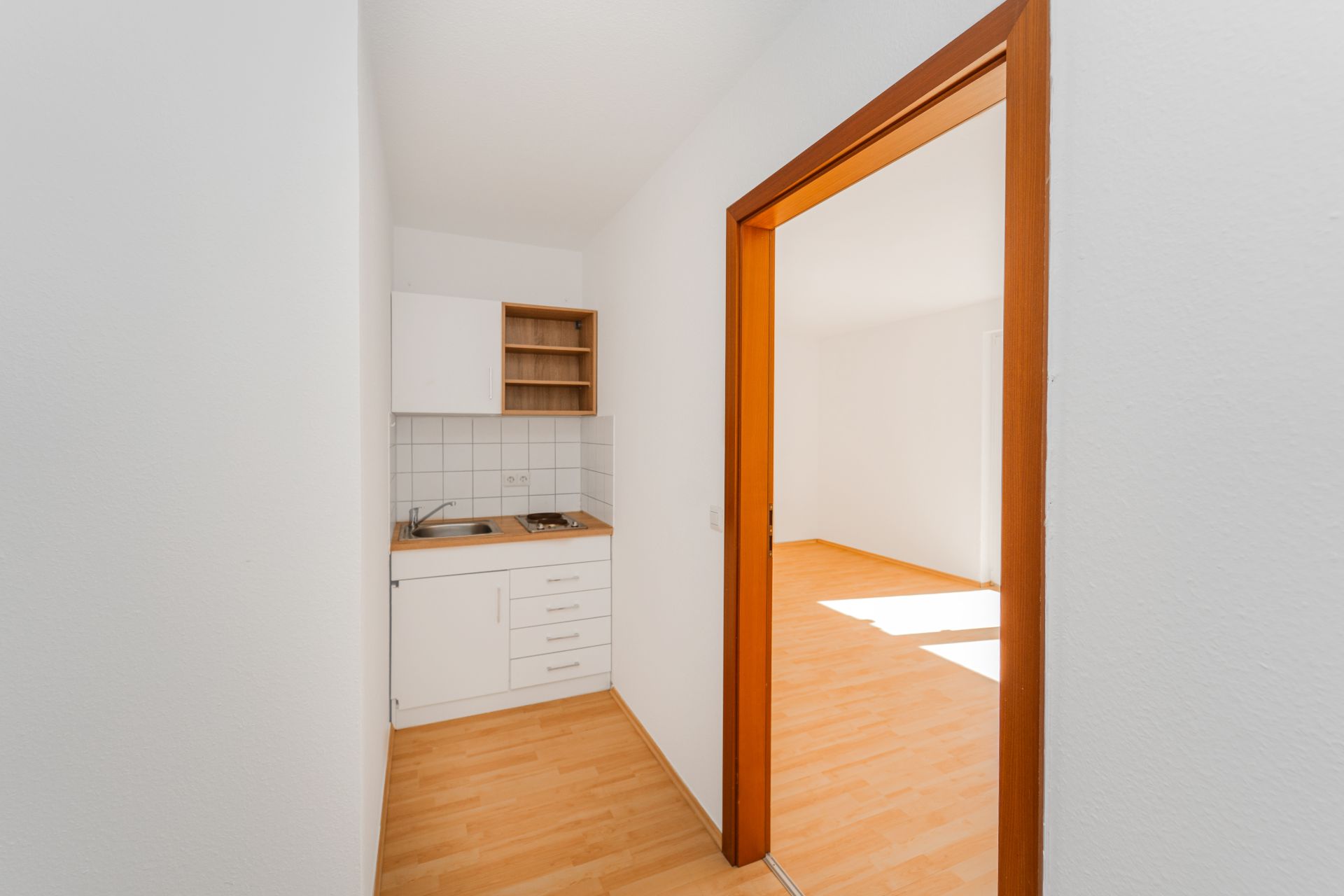Kleine Küchenzeile mit Spüle und Herd, angrenzend an helles Zimmer mit Holzfußboden.