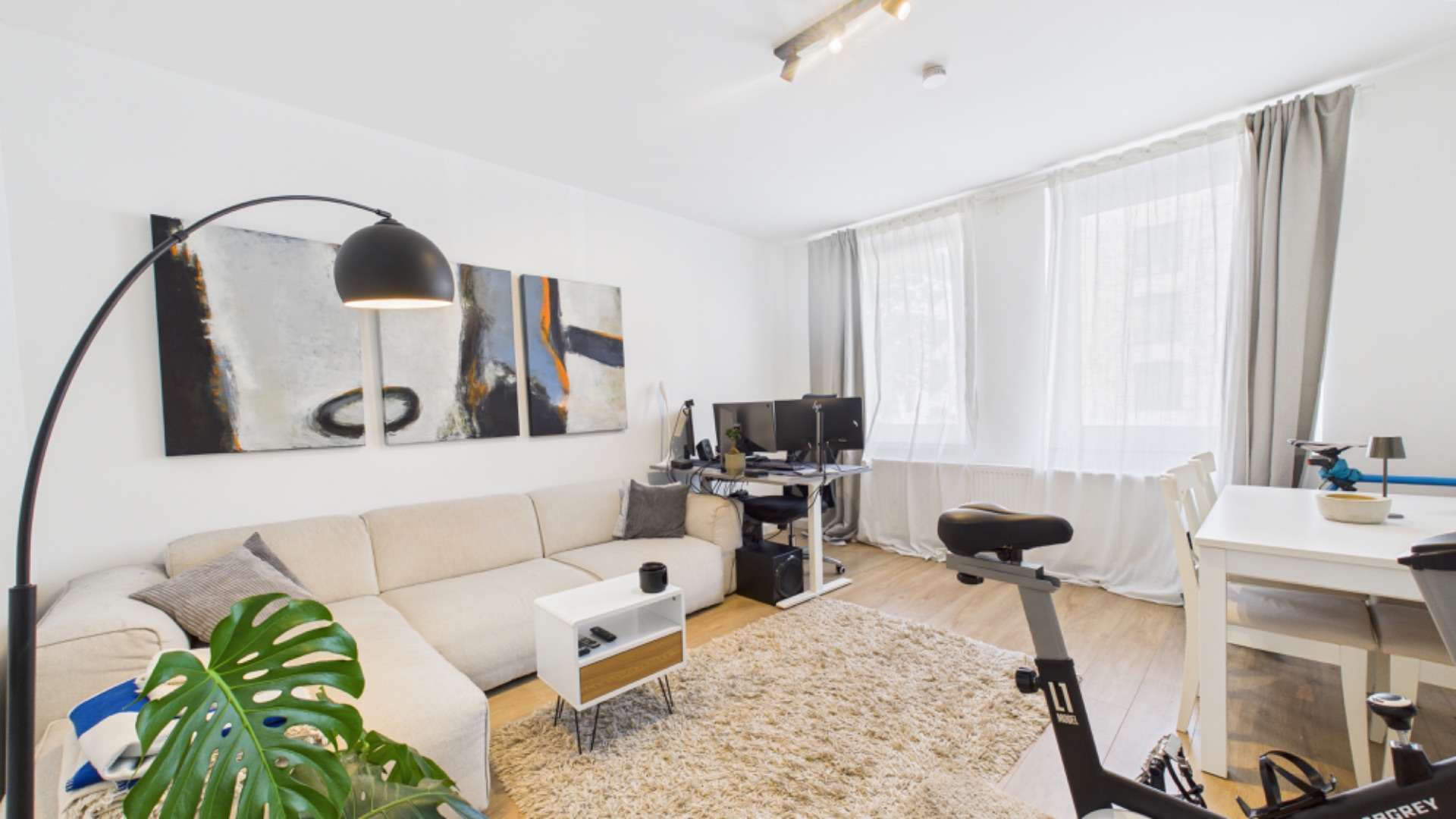 Helles Wohnzimmer mit Ecksofa, Schreibtisch, Heimtrainer und großem Teppich.