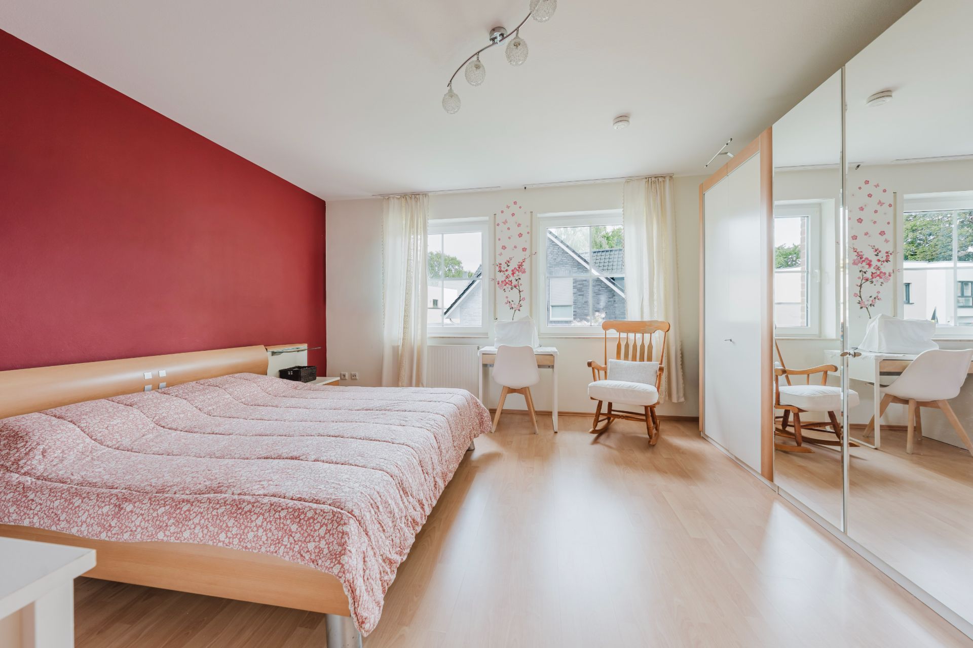 Helles Schlafzimmer mit Doppelbett, roter Wand, Spiegel-Schrank und großen Fenstern.