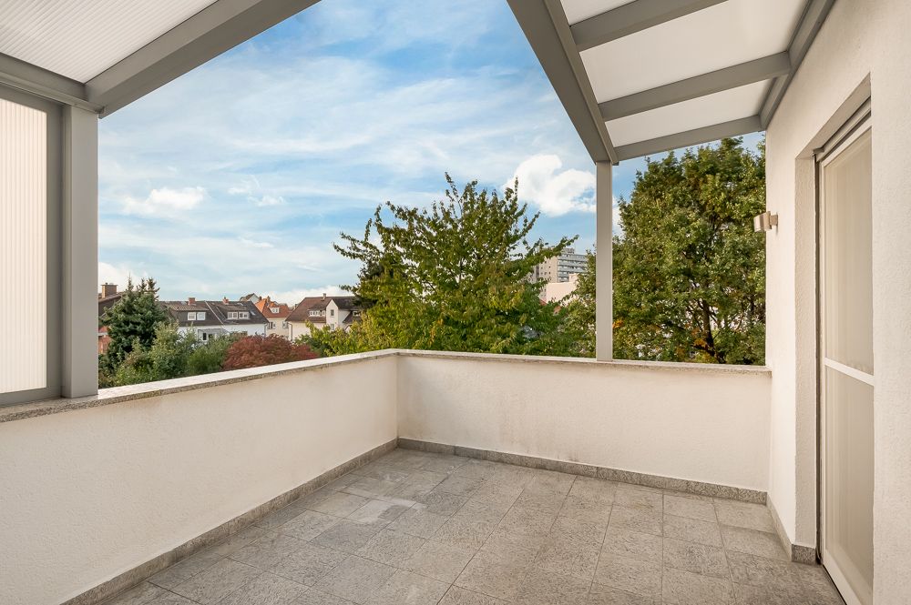 Überdachter Balkon mit Fliesenboden und Ausblick auf Bäume und Wohnhäuser.