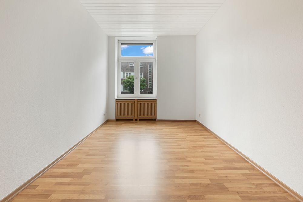 Leerer Raum mit Parkettboden, weiß gestrichenen Wänden und Fenster mit Blick nach draußen.