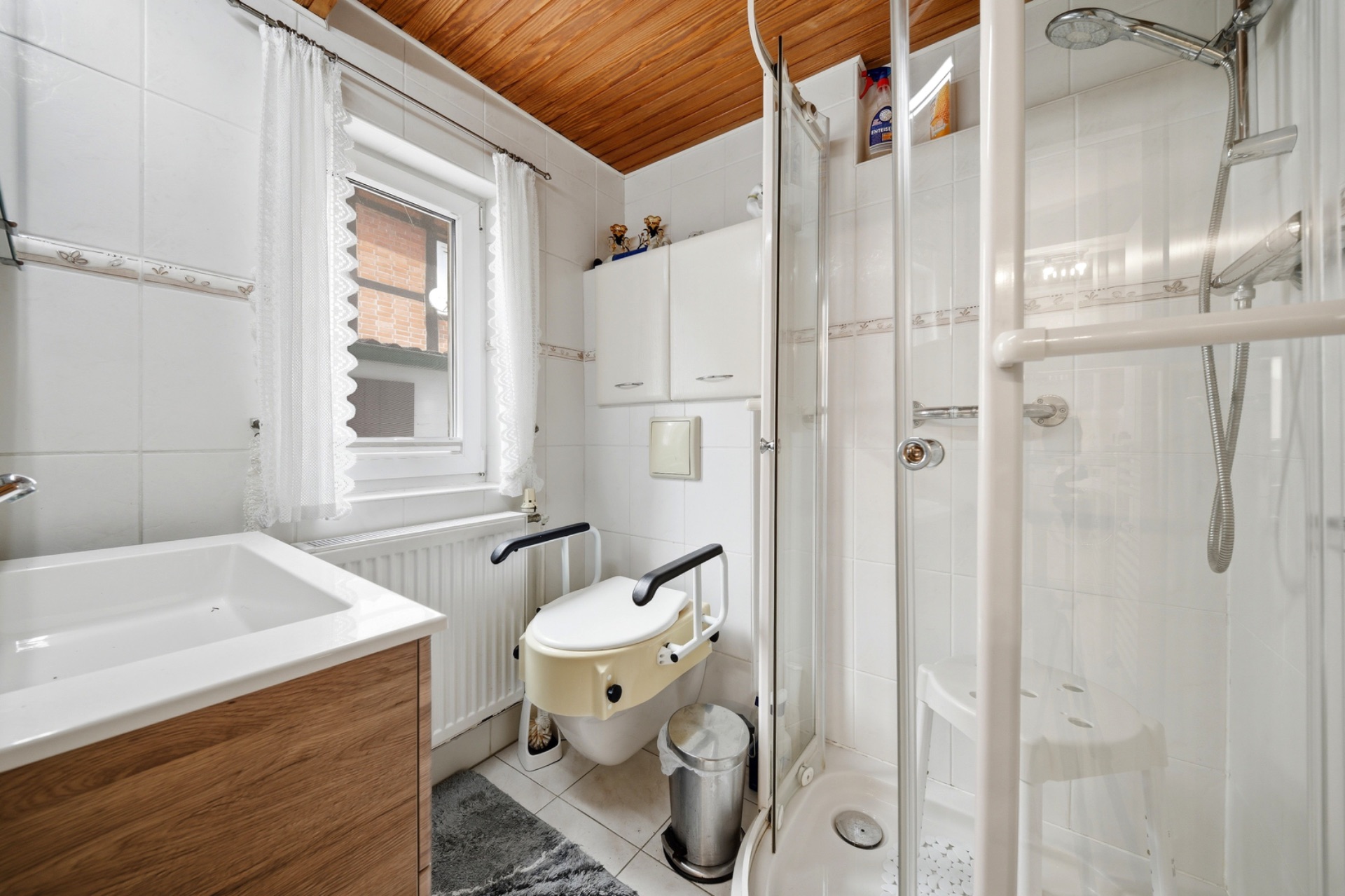 Badezimmer mit Waschbecken, Wandtoilette mit Haltegriffen und Dusche, Fenster mit Vorhang.