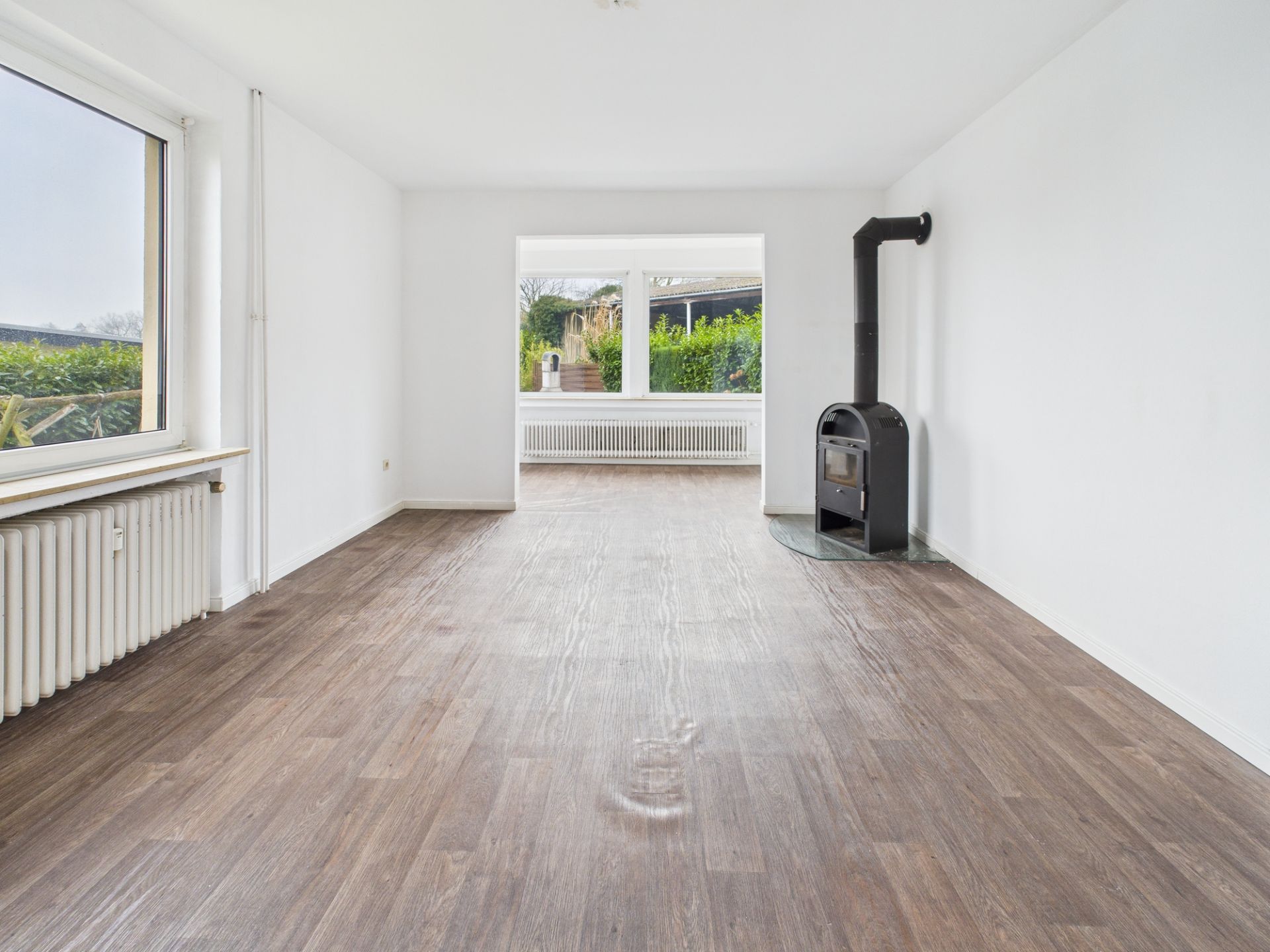 Heller Wohnraum mit Holzfußboden, Kaminofen und großen Fenstern, Blick in den Garten.