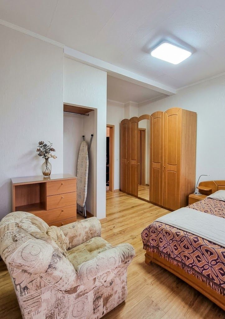 Helles Schlafzimmer mit Sessel, Bett, Holzschränken, Kommode und Bügelbrett.