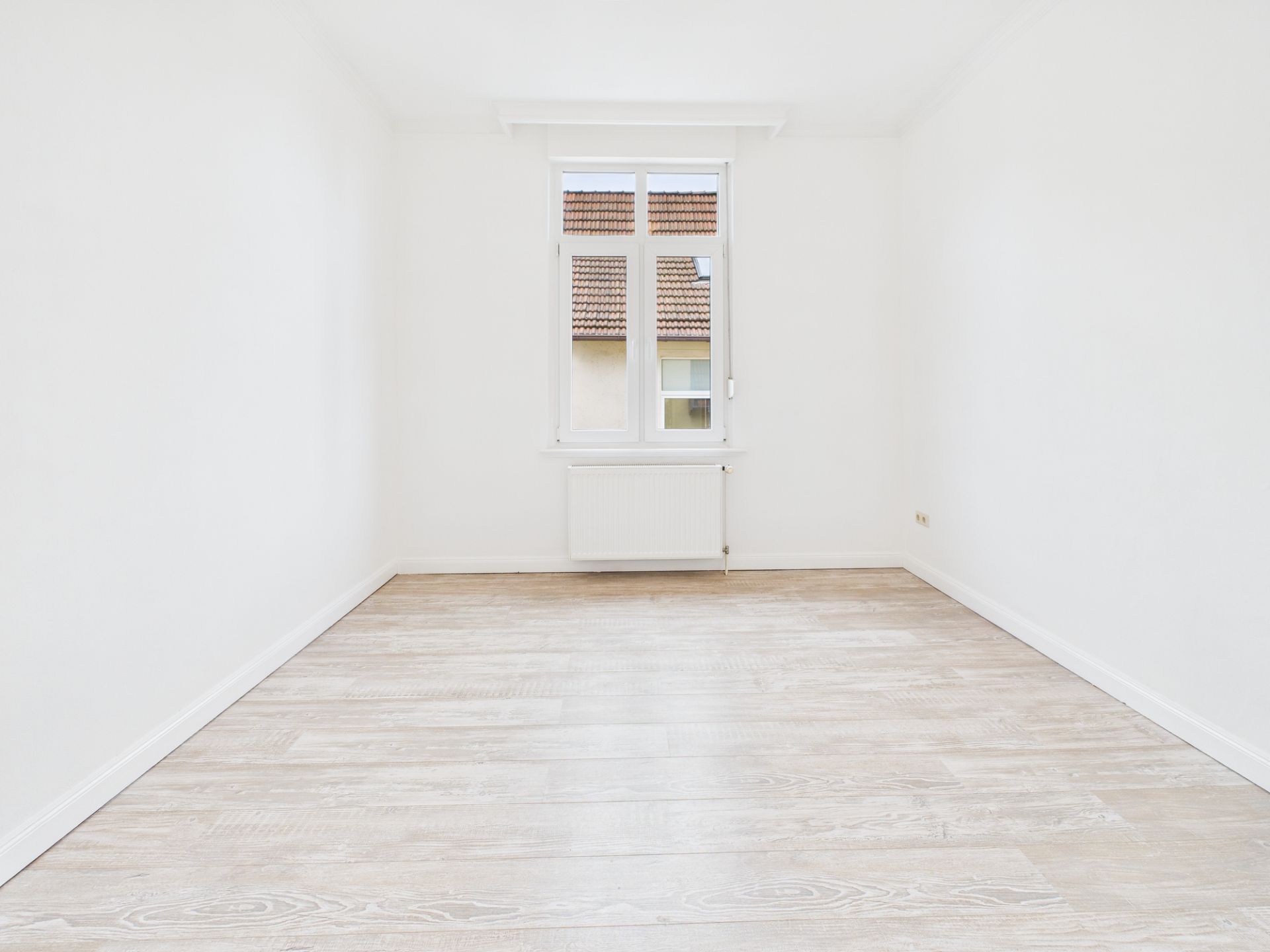 Leerer, heller Raum mit weißem Wänden, Holzfußboden und einem Fenster mit Blick auf Dächer.