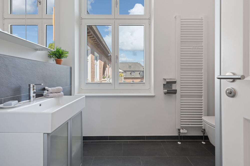 Helles Badezimmer mit Waschbecken, Handtüchern, Heizung, WC und großem Fenster.