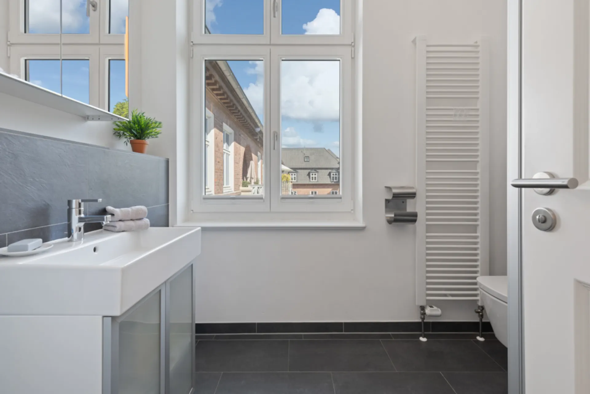 Helles Badezimmer mit Waschbecken, Handtüchern, Heizung, WC und großem Fenster.