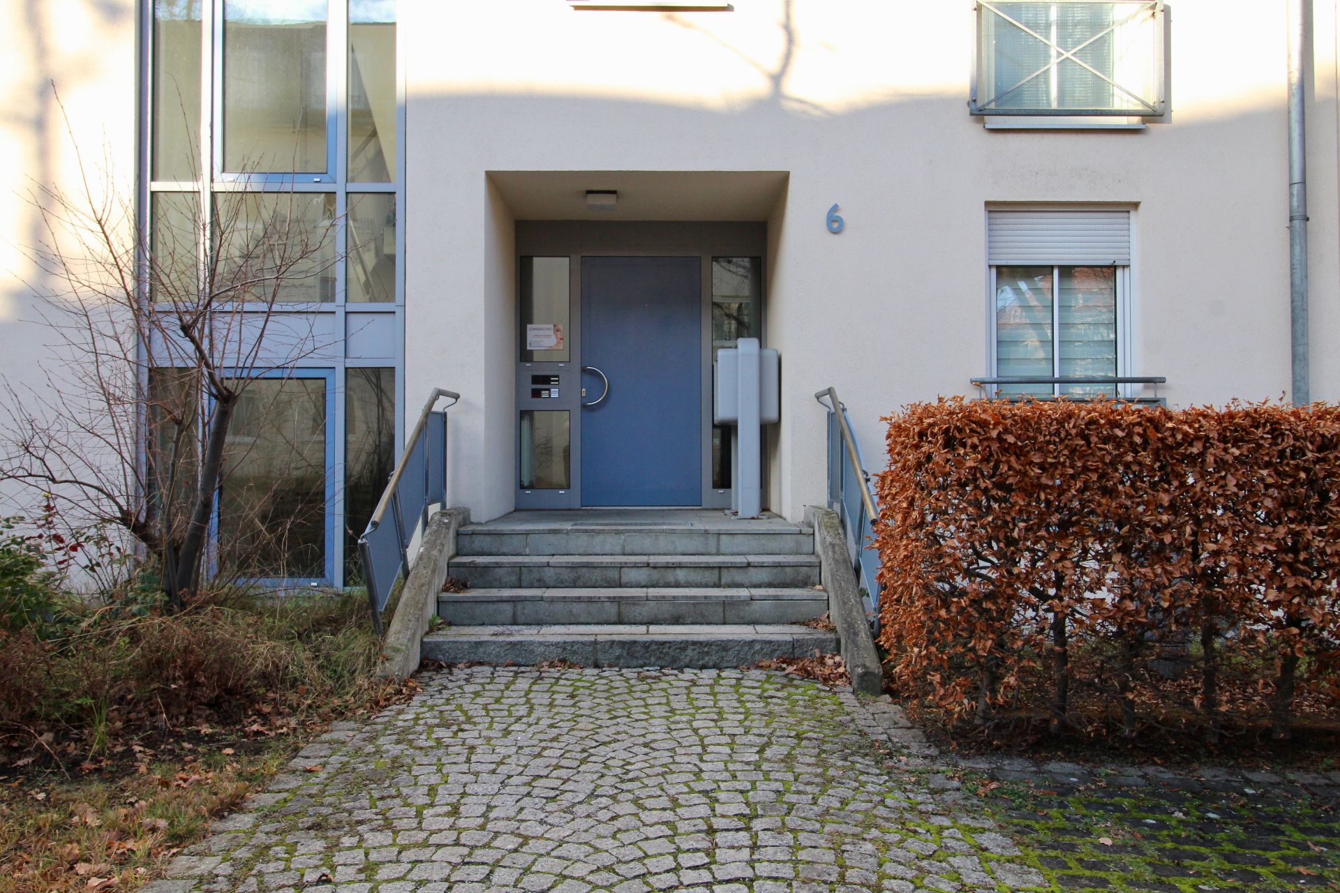 Eingang eines Wohnhauses mit Treppe, blauer Tür und Briefkästen, umgeben von Pflanzen.
