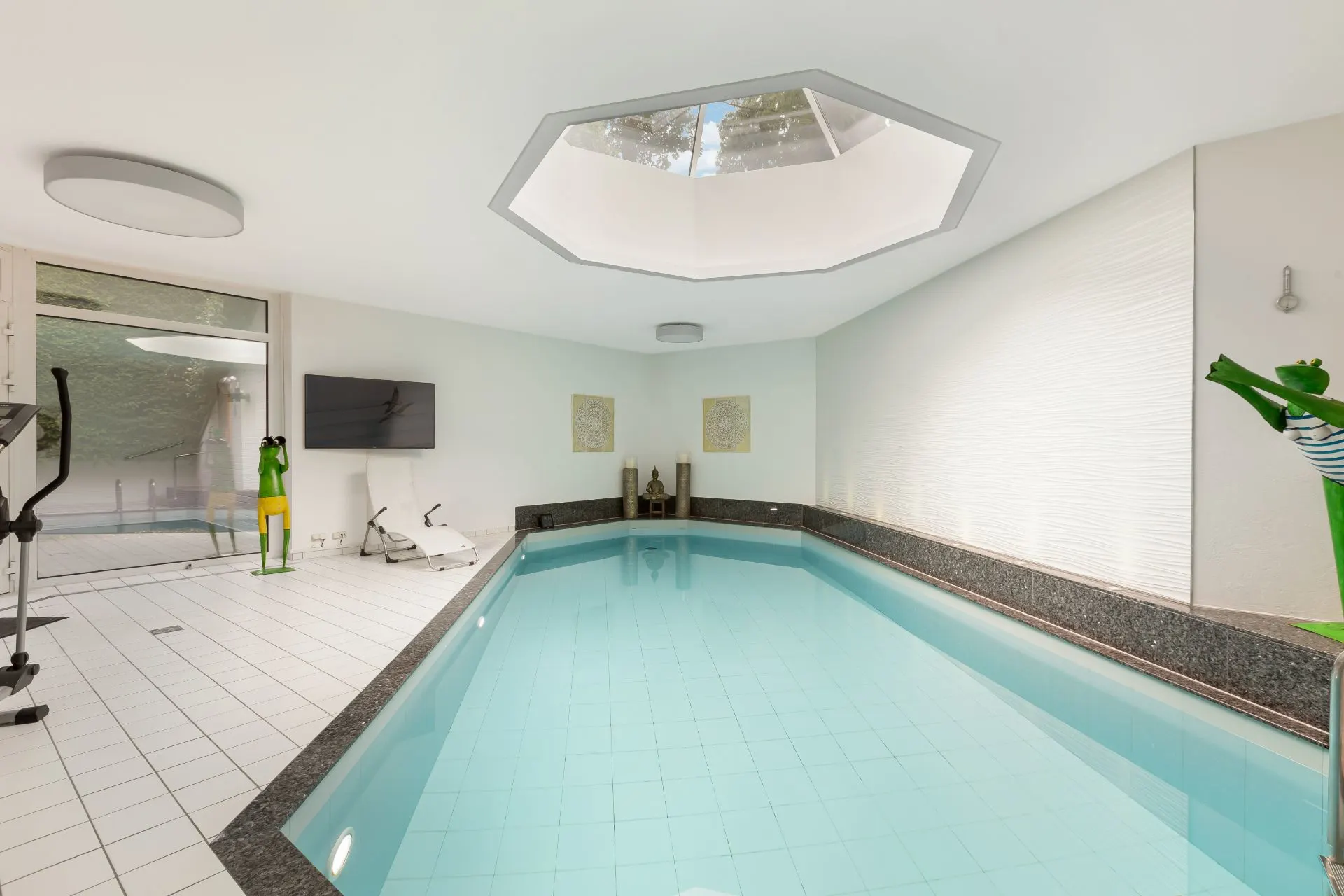 Heller Innenpool mit Fitnessgeräten und großem Deckenfenster in einem Raum mit weißen Fliesen.
