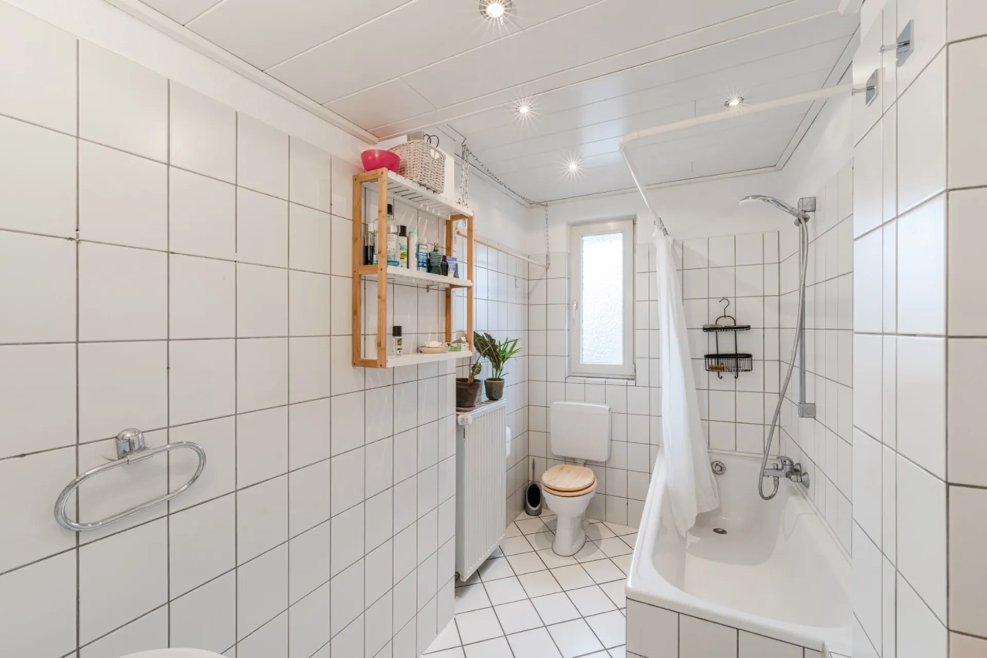 Helles Badezimmer mit Badewanne, WC, Regal und weiß gefliesten Wänden.