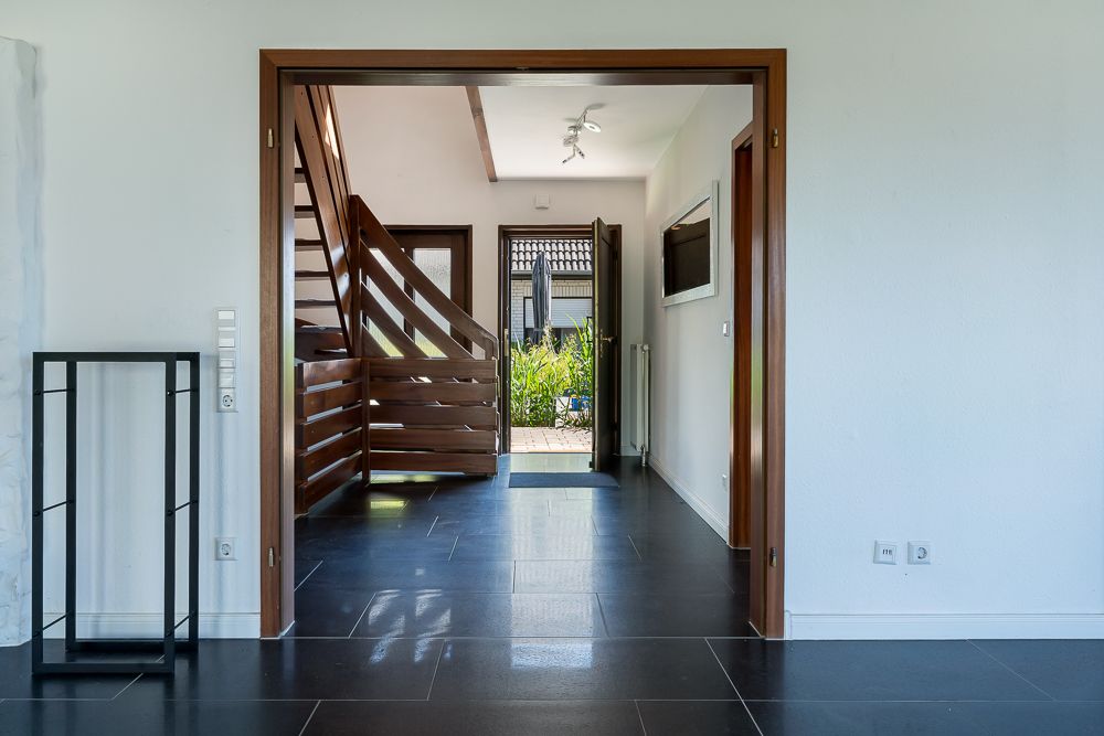 Flur mit schwarzem Fliesenboden, Holztreppe und offen stehender Tür zum Garten.