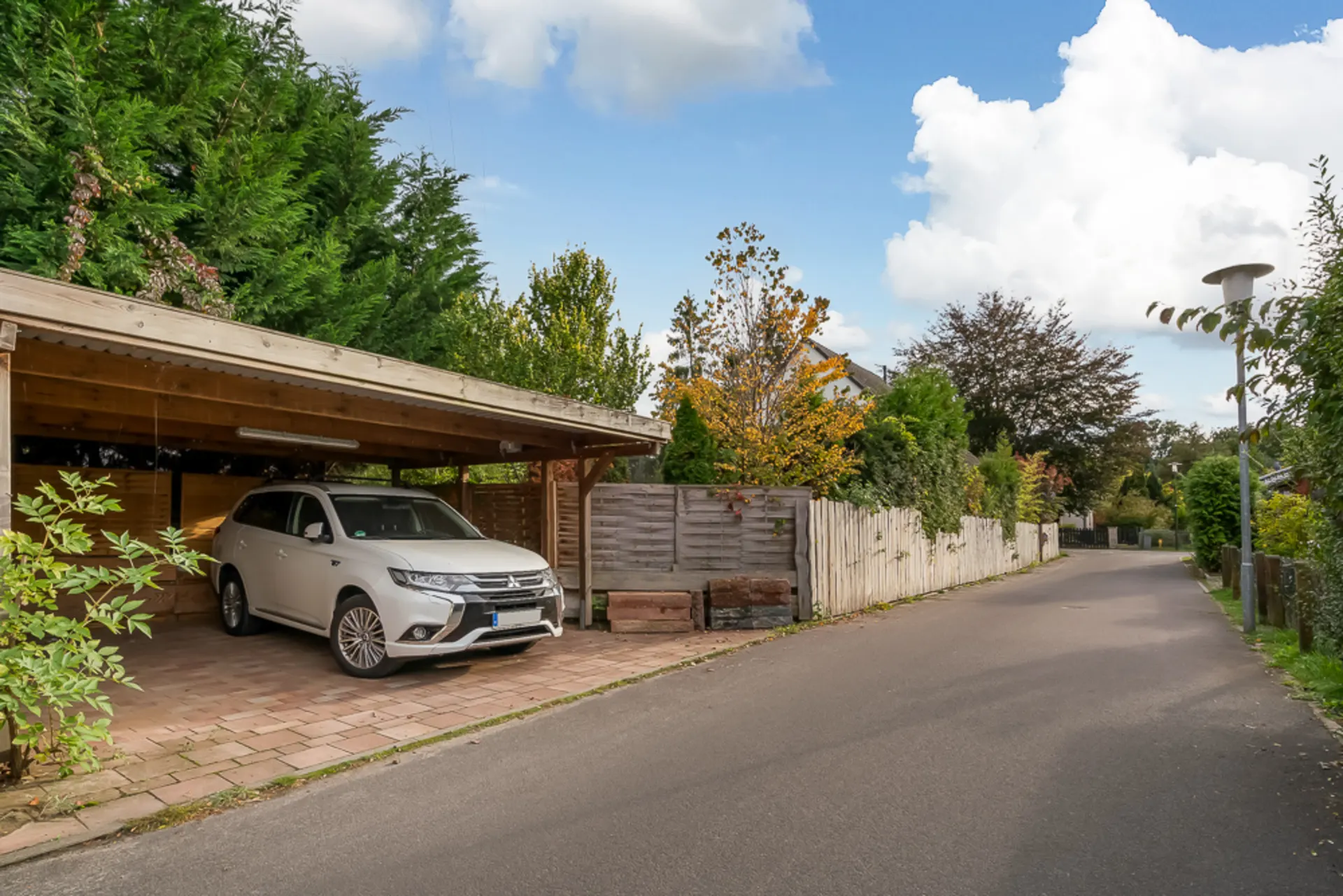 Weißes Auto unter einem Carport neben einer ruhigen Wohnstraße mit Bäumen und Holzzaun.