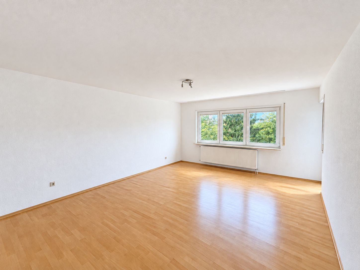 Helles, leeres Zimmer mit Holzfußboden und drei Fenstern, durch die Bäume zu sehen sind.