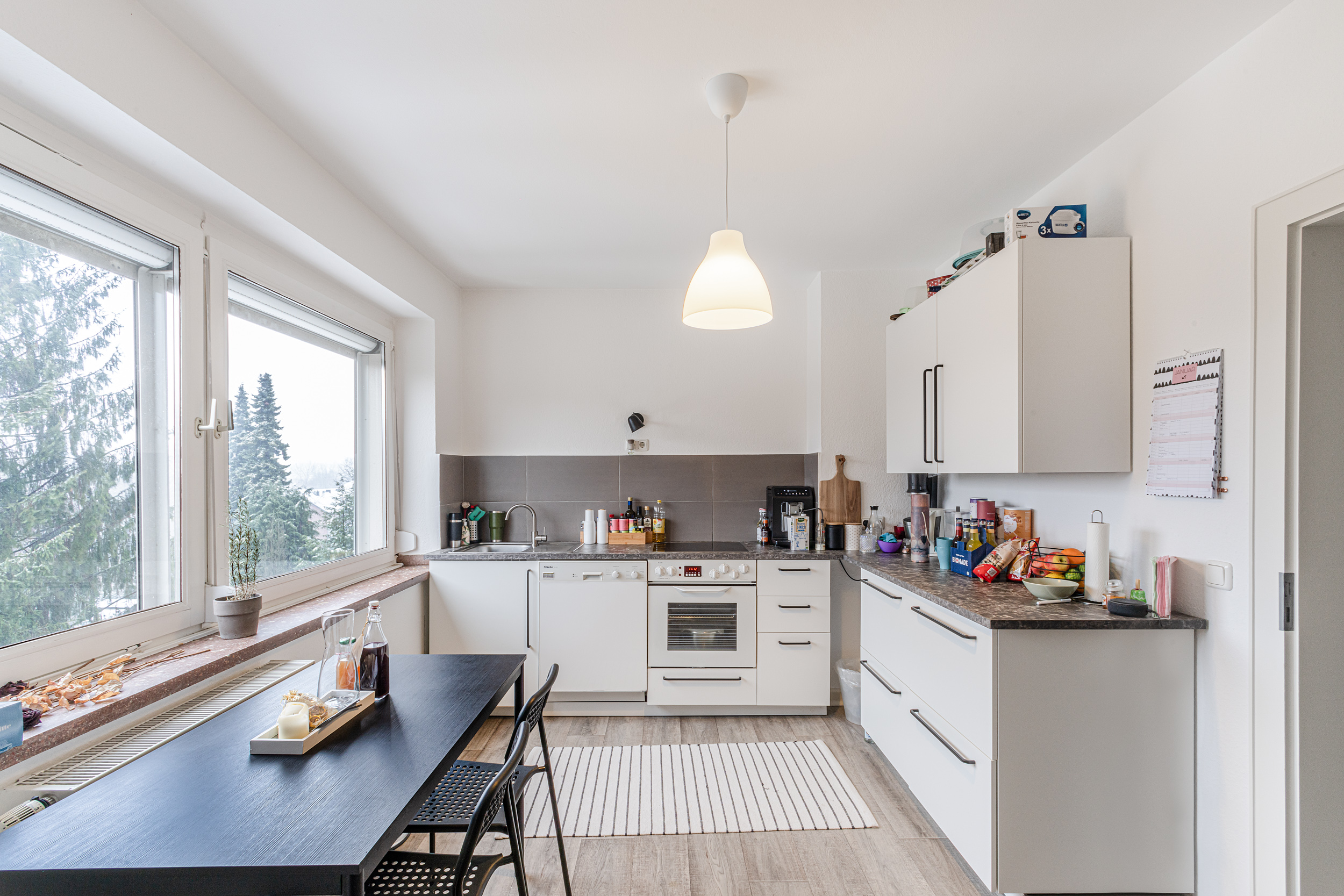 Helle Küche mit weißen Schränken, Esstisch, großen Fenstern und Hängelampe.