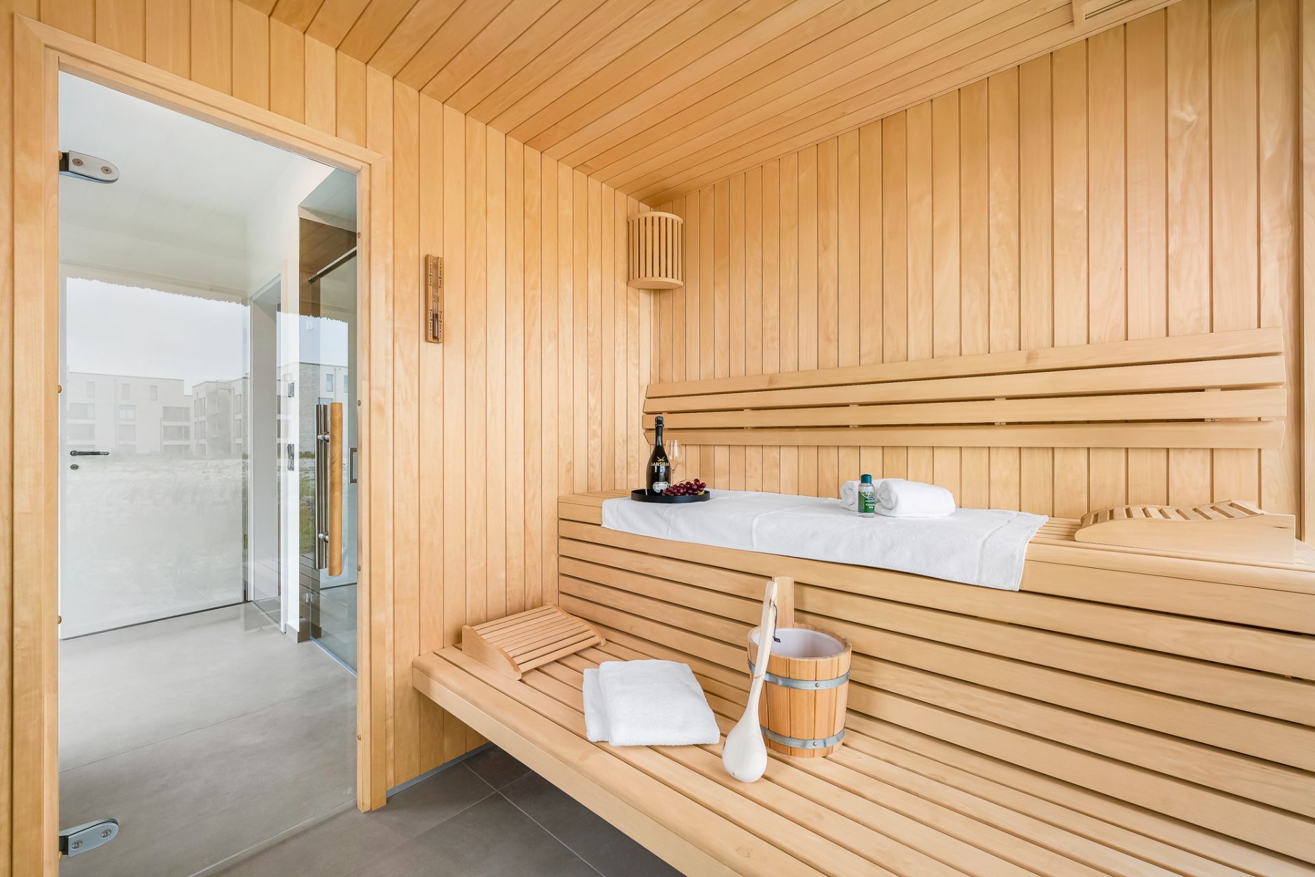 Helle Sauna aus Holz mit Handtuch, Saunaeimer, Weinflasche und Gläsern auf der Bank.