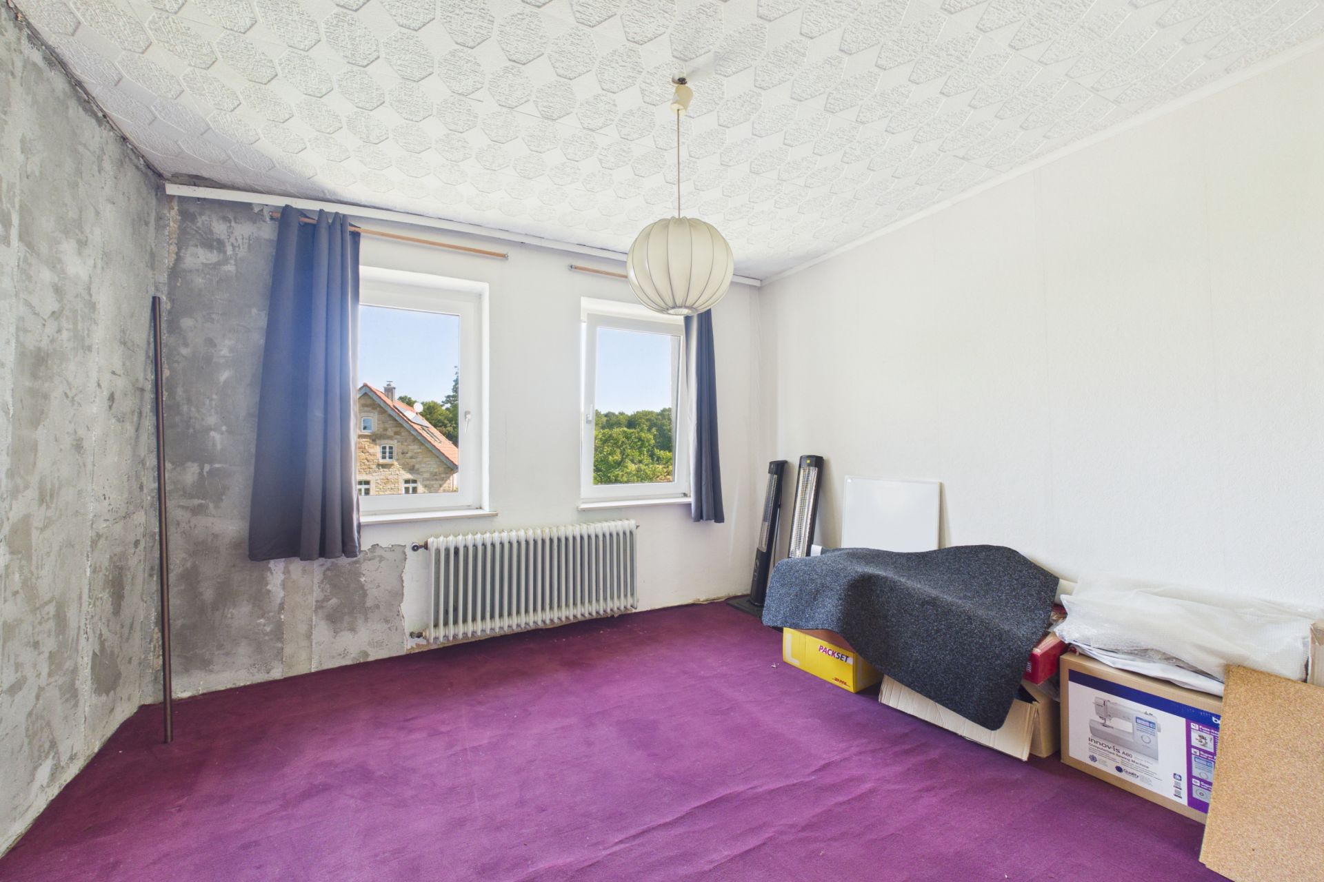 Zimmer mit lila Teppich, zwei Fenstern, Heizung und einigen Kartons auf dem Boden.
