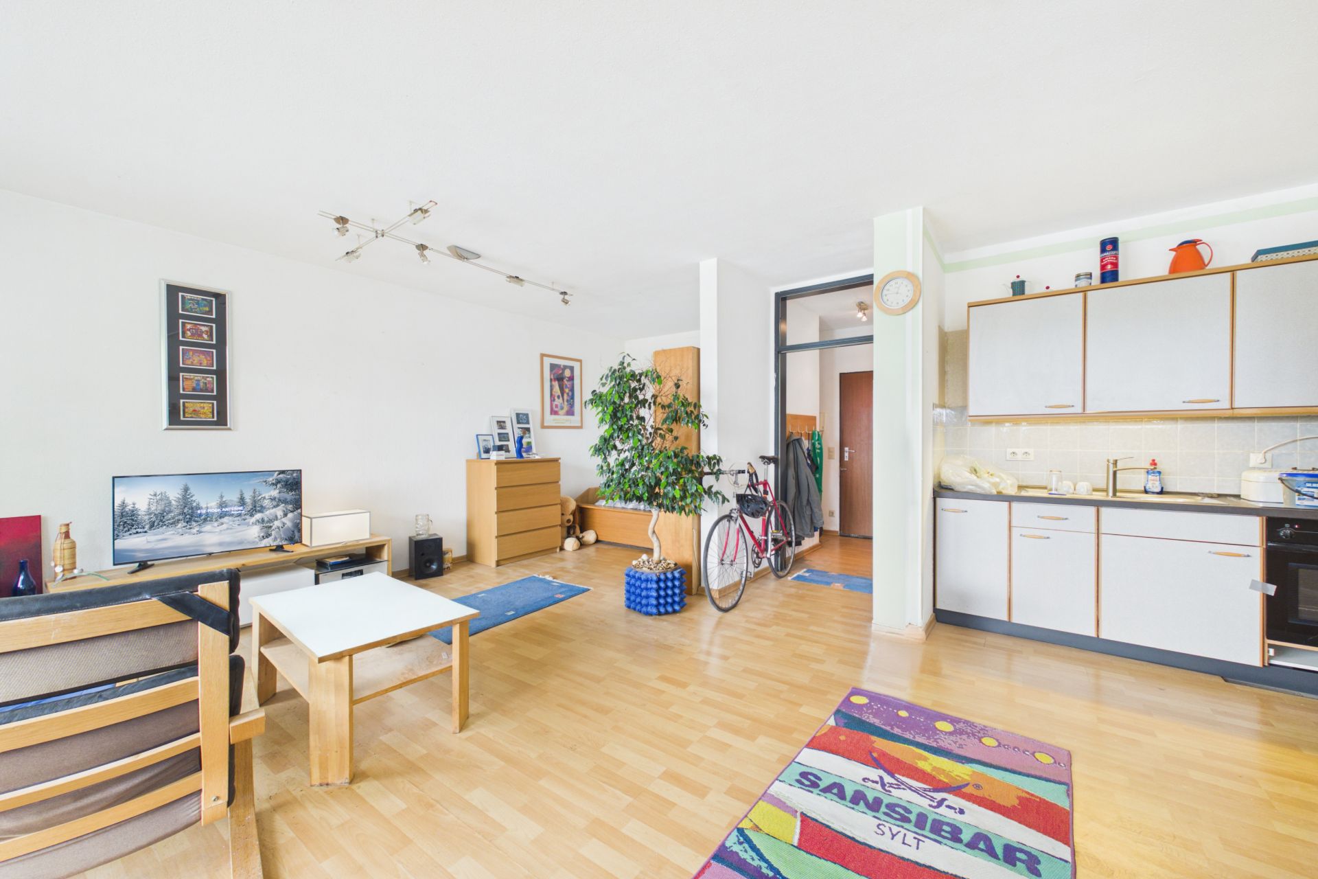 Helles Wohnzimmer mit Holzboden, offener Küche, Möbeln, Fernseher, Pflanze und Fahrrad.