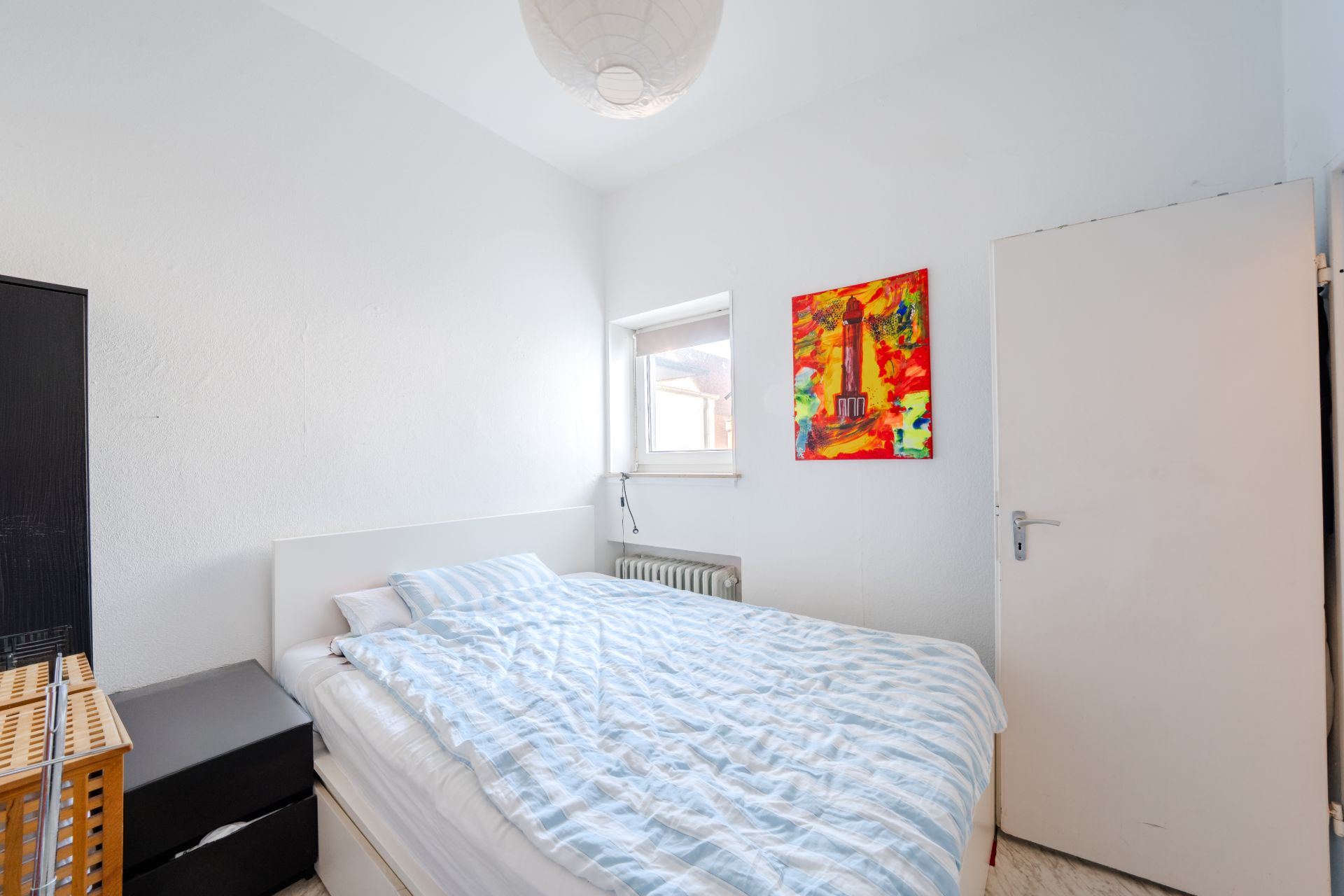 Helles Schlafzimmer mit Bett, Fenster, bunter Wandkunst und weißer Tür.