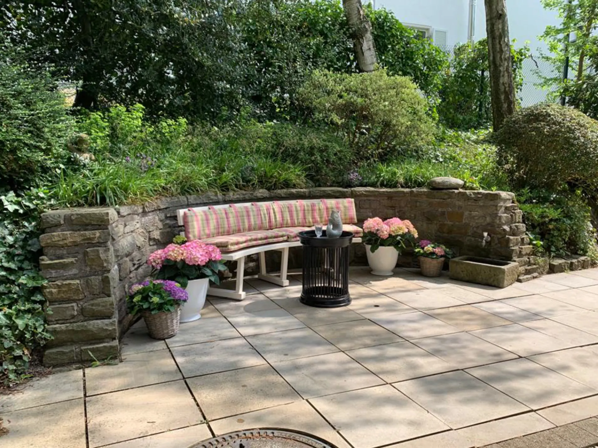 Gepflasterte Terrasse mit Sitzbank, Blumen in Töpfen und kleinem Tisch vor einer Steinmauer.