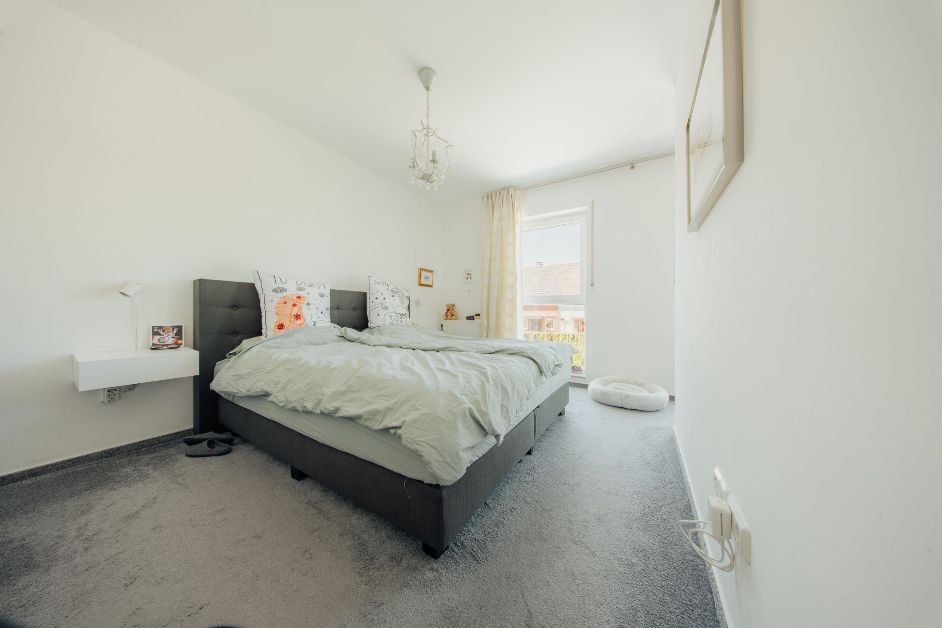 Helles Schlafzimmer mit Doppelbett, Nachttisch und Fenster mit Vorhang.