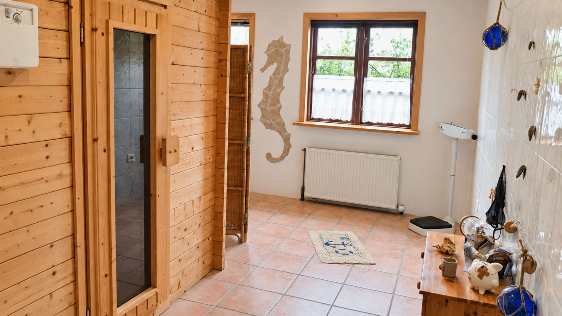Heller Raum mit Holzsauna, Fliesenboden, Fenster, Heizkörper und Wanddeko in Form eines Seepferdchens.