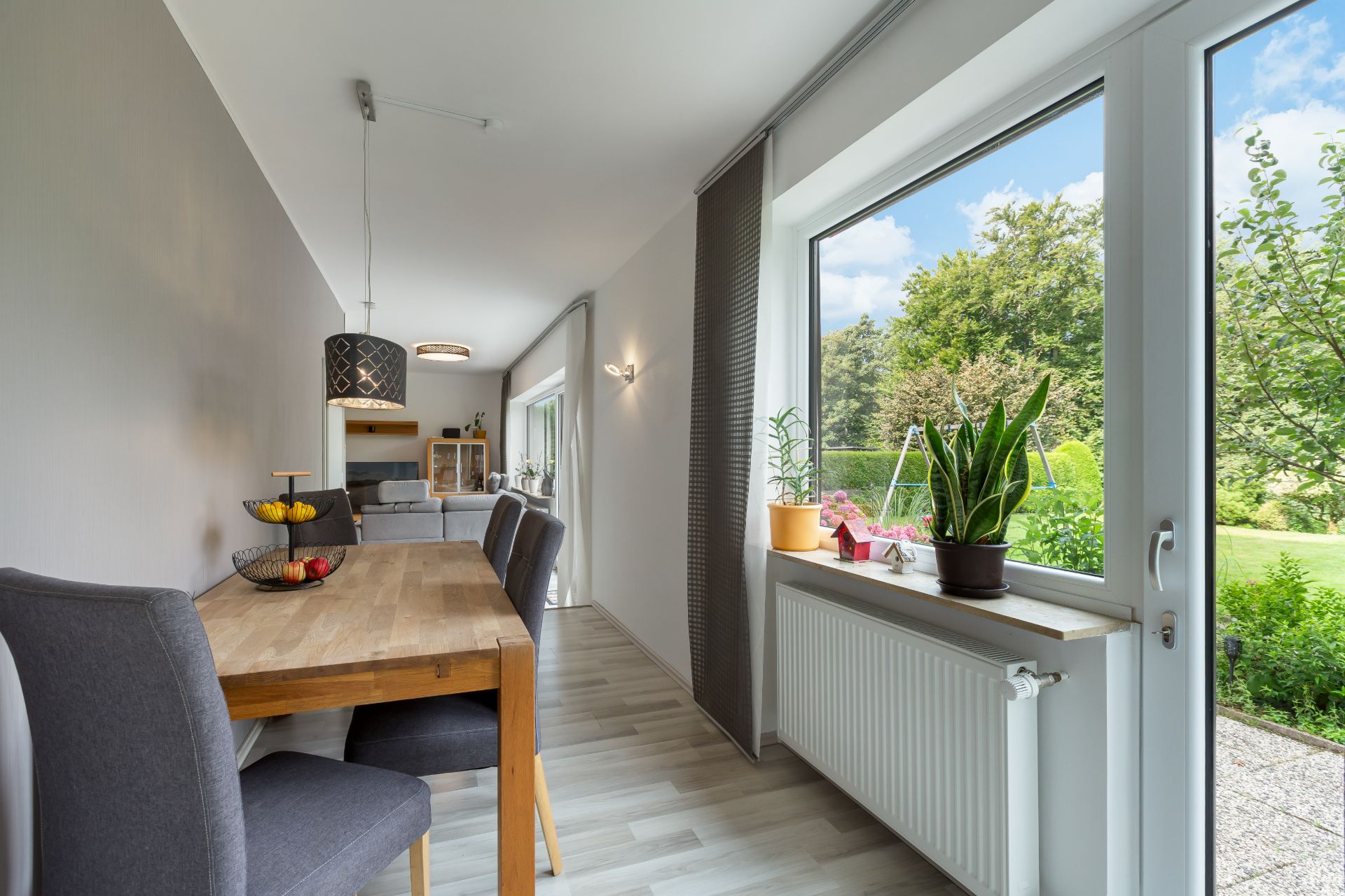 Essbereich mit Holztisch und grauen Stühlen neben großem Fenster, Blick in den Garten.